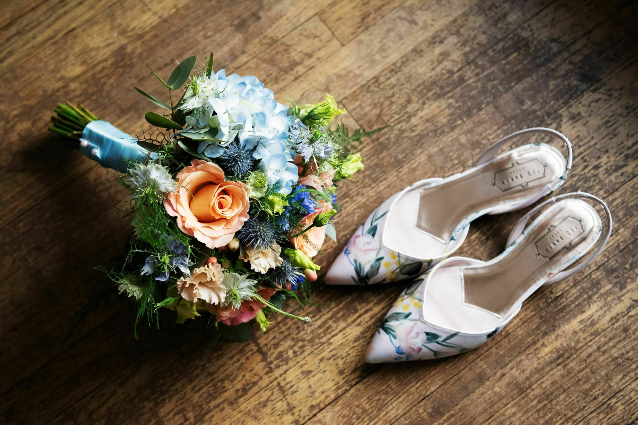 A bouquet of flowers with blue and peach roses next to a pair of floral patterned high-heeled shoes on a wooden floor, captured beautifully by a wedding photographer at Deer Park Country House wedding.