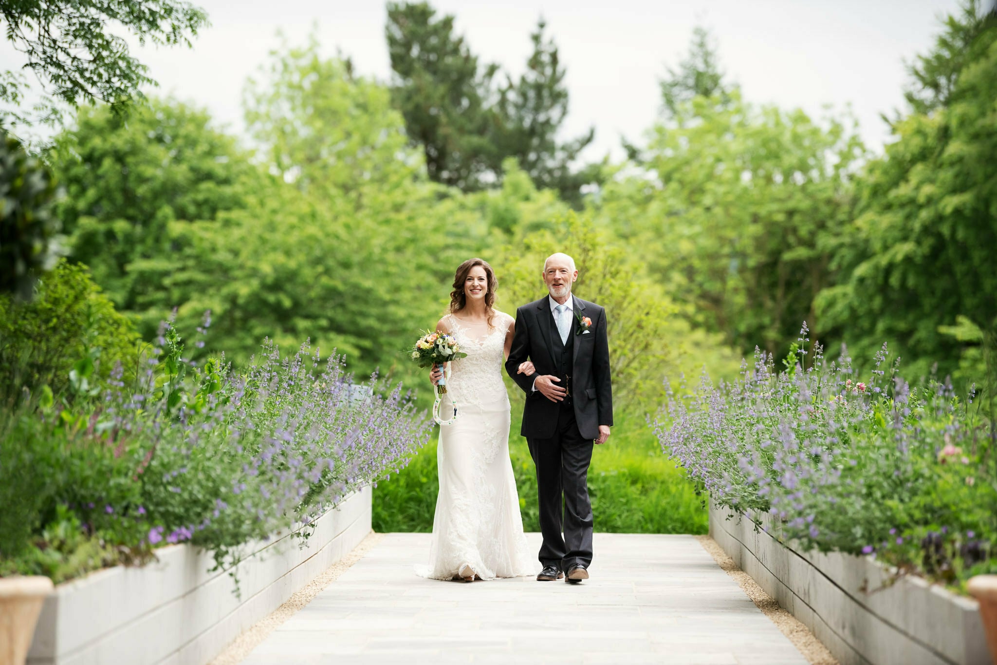 A bride in a white gown and a man in a black suit walk down an outdoor path lined with purple flowers and greenery, capturing the perfect moment for their wedding photography at the enchanting Deer Park Country House in Devon.