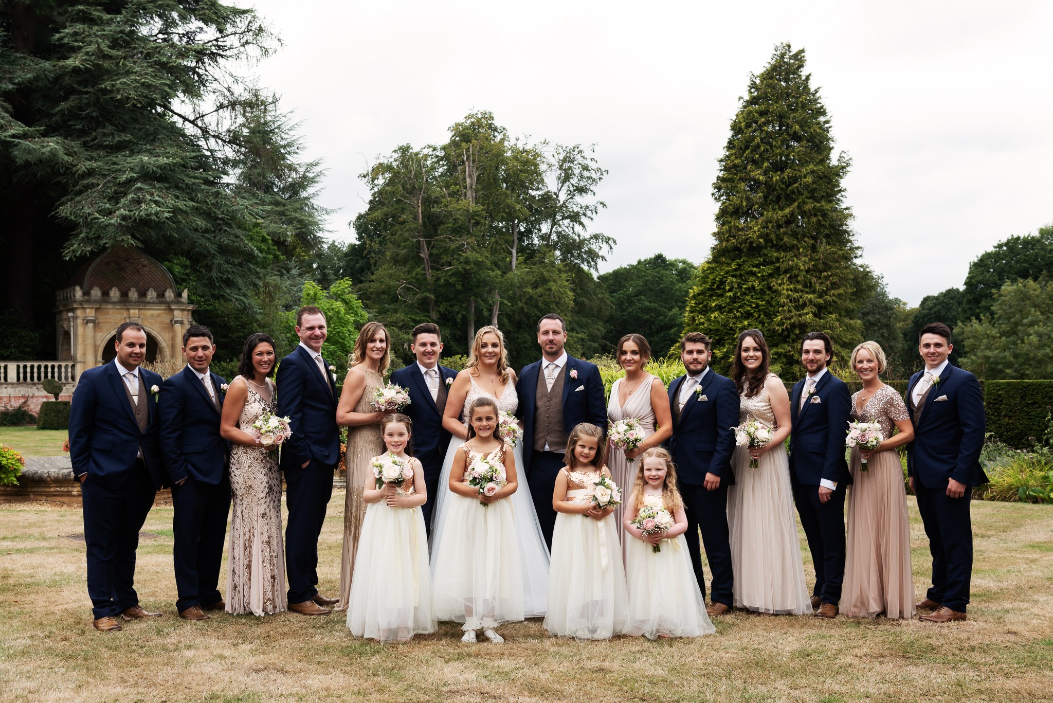 A wedding party stands outdoors at Tylney Hall, featuring a bride, groom, bridesmaids, groomsmen, and flower girls in formal attire, with trees and grass in the background.