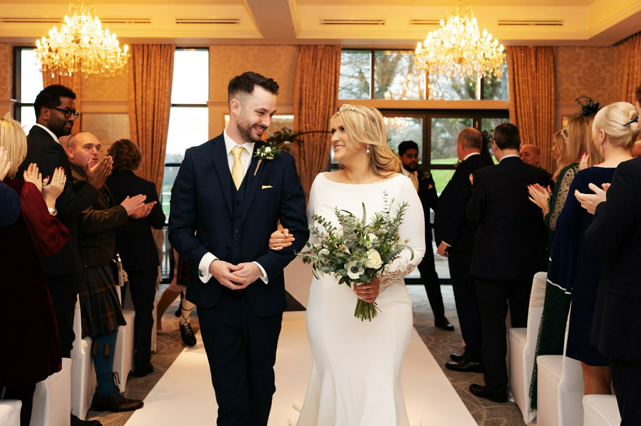 A bride and groom walk down the aisle at their Latimer Estate Wedding, smiling at each other, while guests on both sides clap and watch.