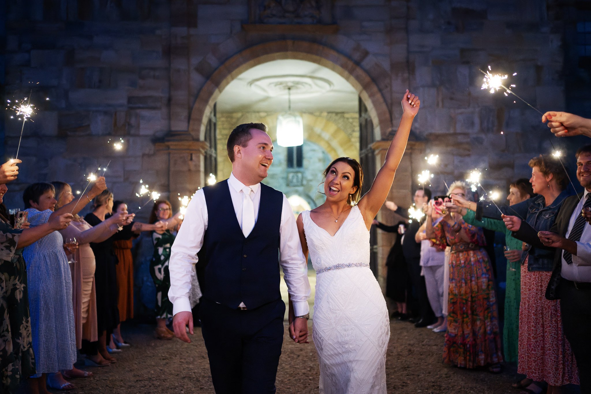 The bride gives a big cheer as she walks with her groom through a channel of her wedding guests waving sparklers at the end of the wedding.