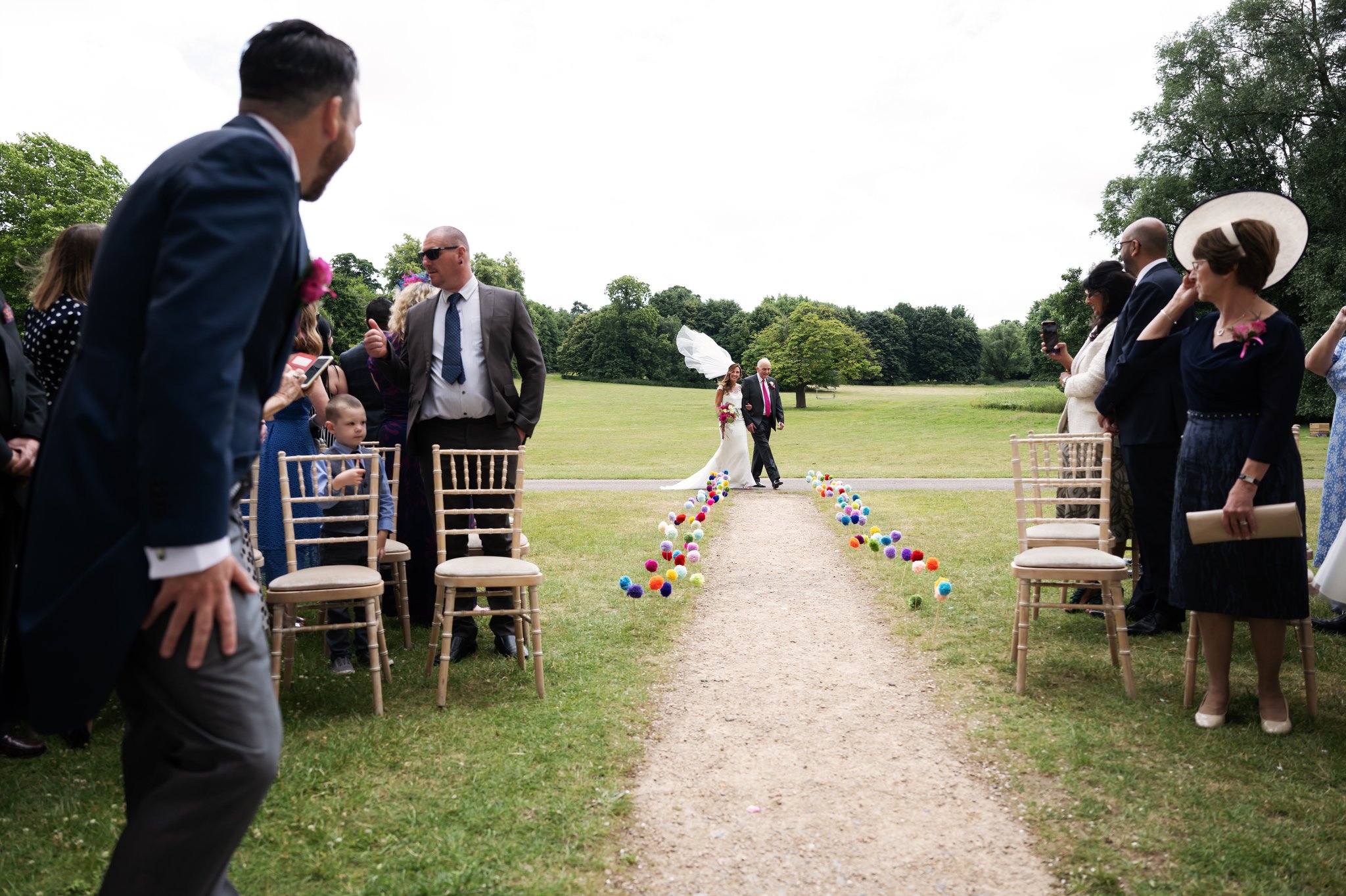 The Brides Veil floats up high as the Bride starts to walk down the aisle at the beginning of an outdoor wedding ceremony on a windy day at Hitchin Priory.