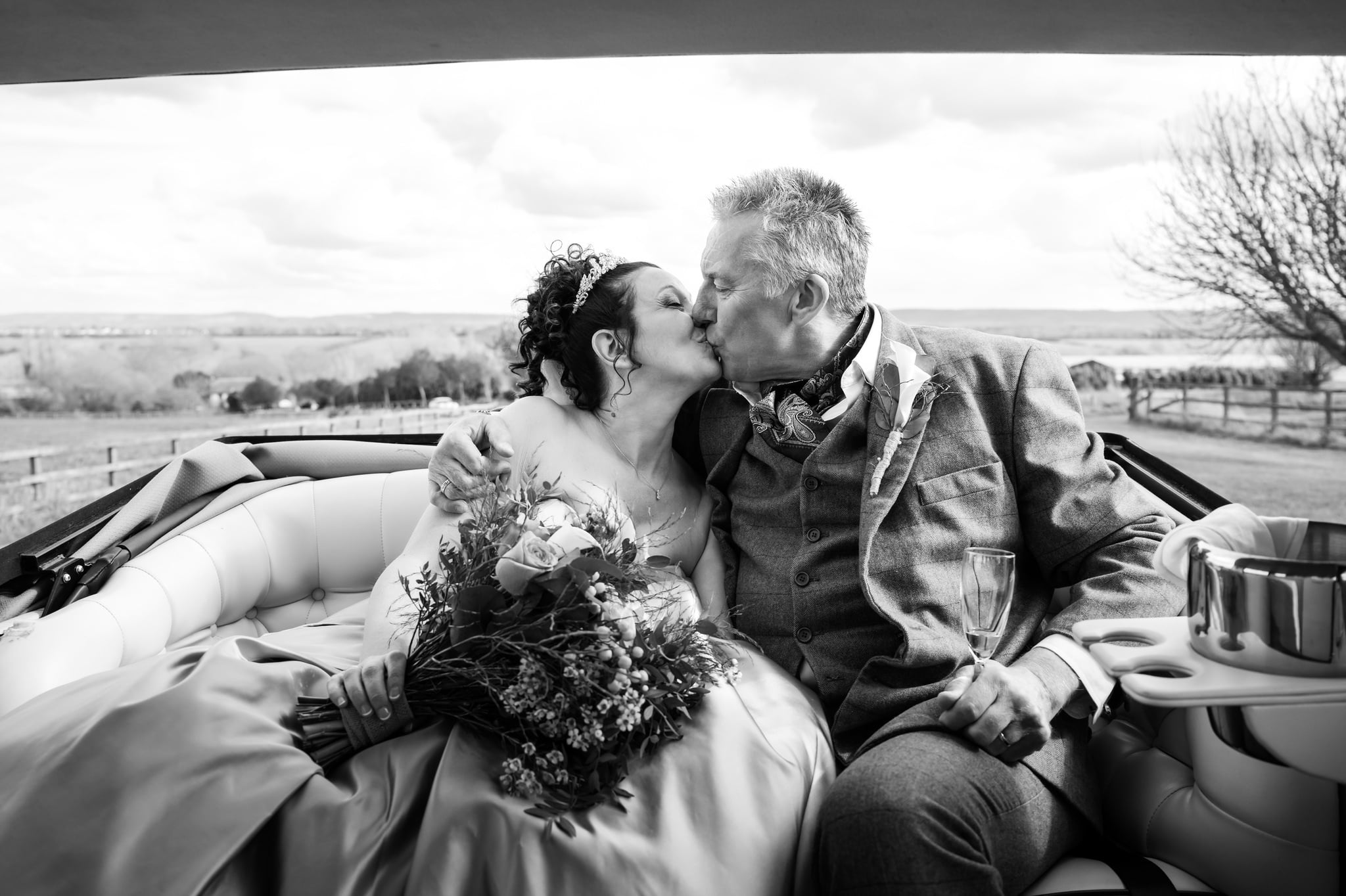 The bride and groom kiss in the back of their wedding car. The Bride is holding her bouquet and the groom is holding a glass of Champagne.
