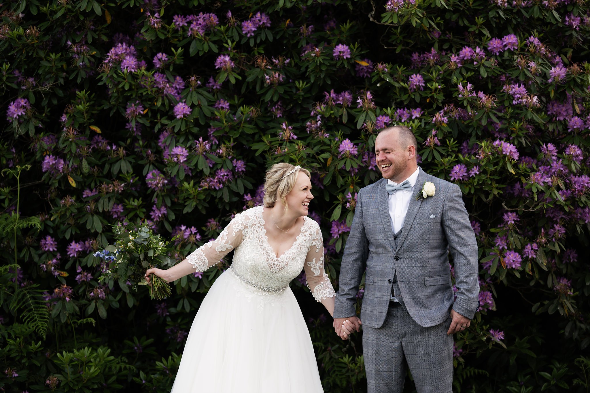 The bride and groom holding hands and laughing in front of a rhododendron bush at Shendish Manor.
