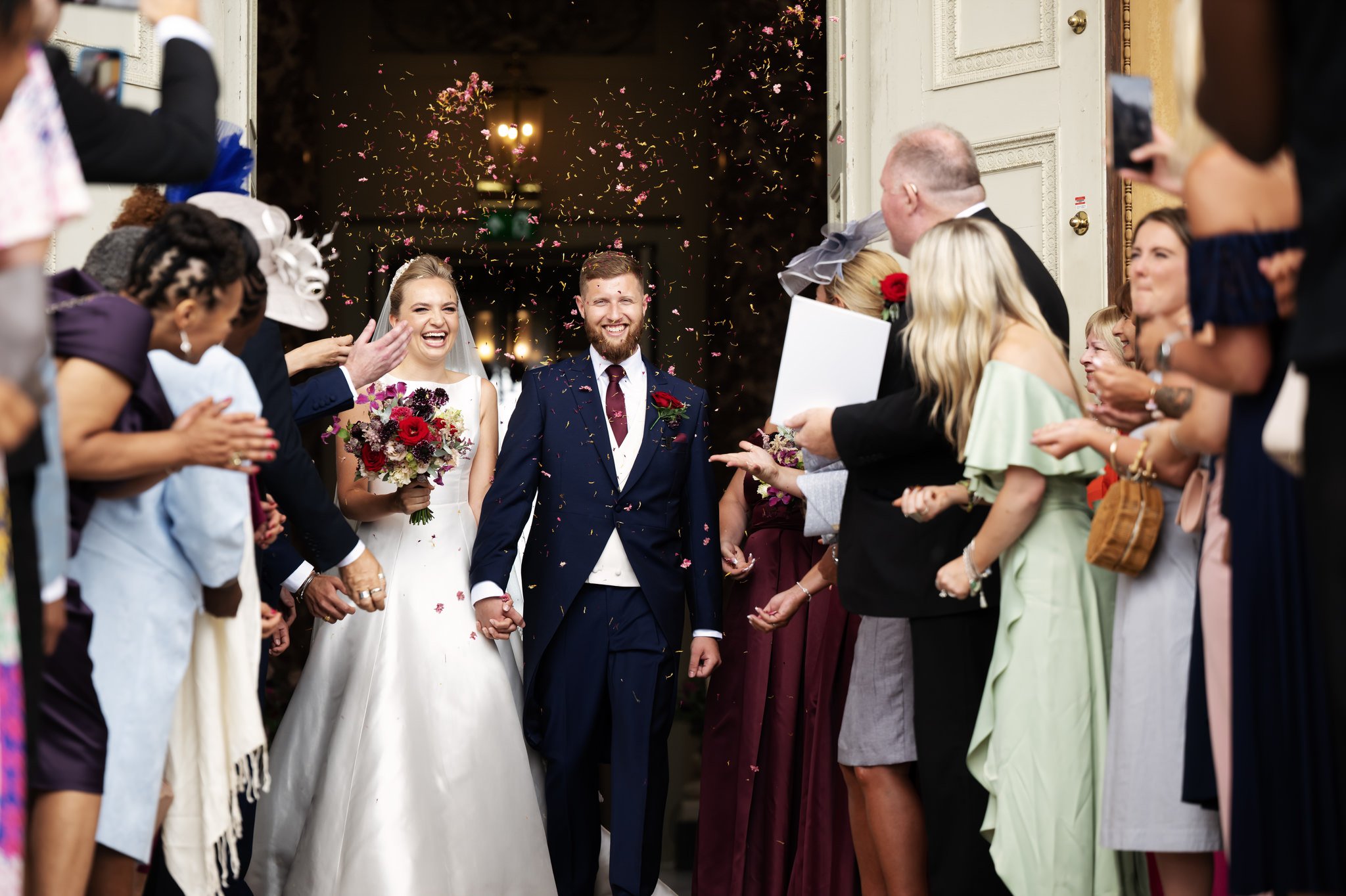 Guests throw confetti over the bride and groom as they walk out of The Marble Hall at the end of their wedding ceremony.