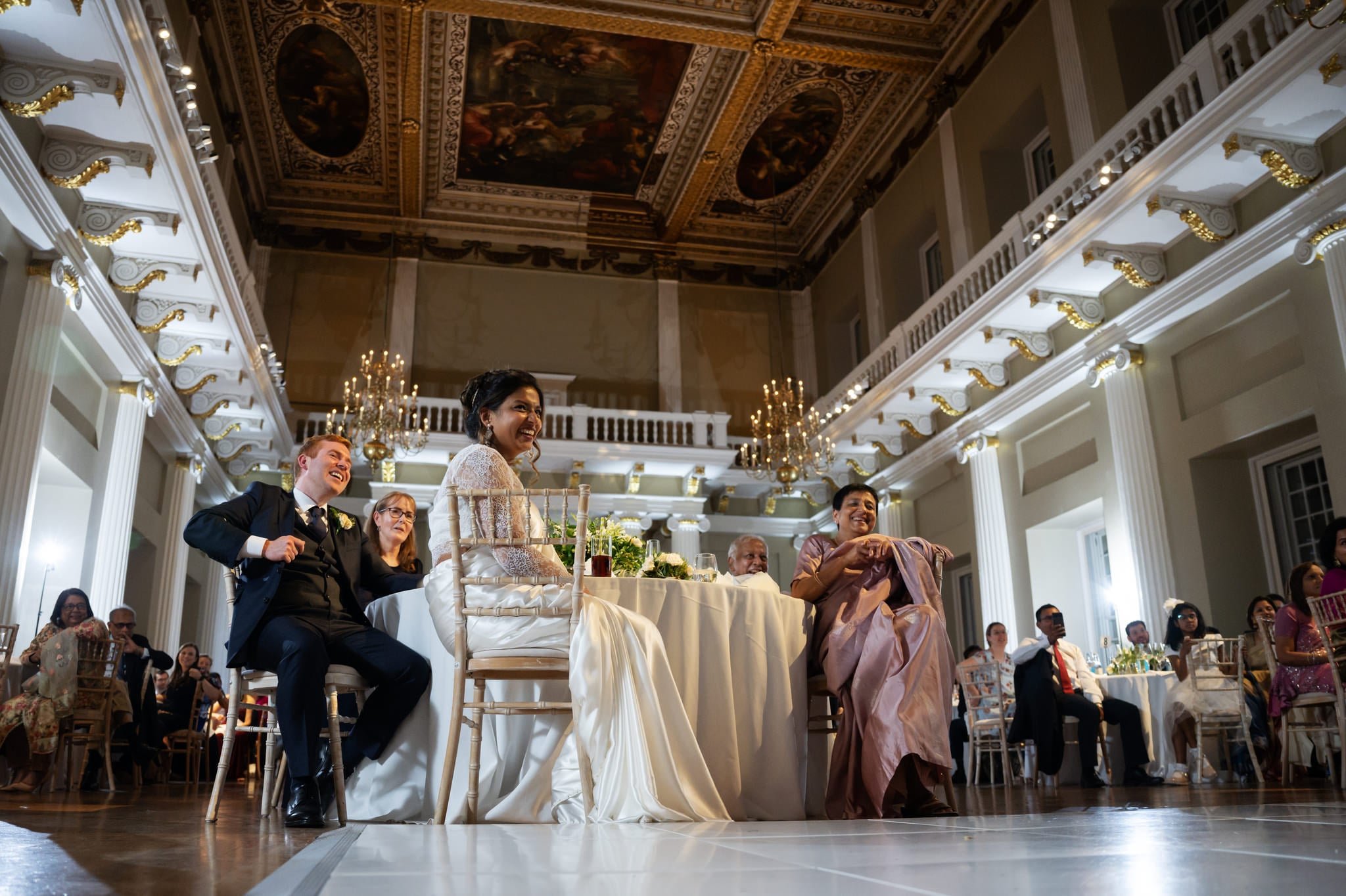 A wide low angle shot of the bride and groom listening to the speeches at a banqueting house wedding.