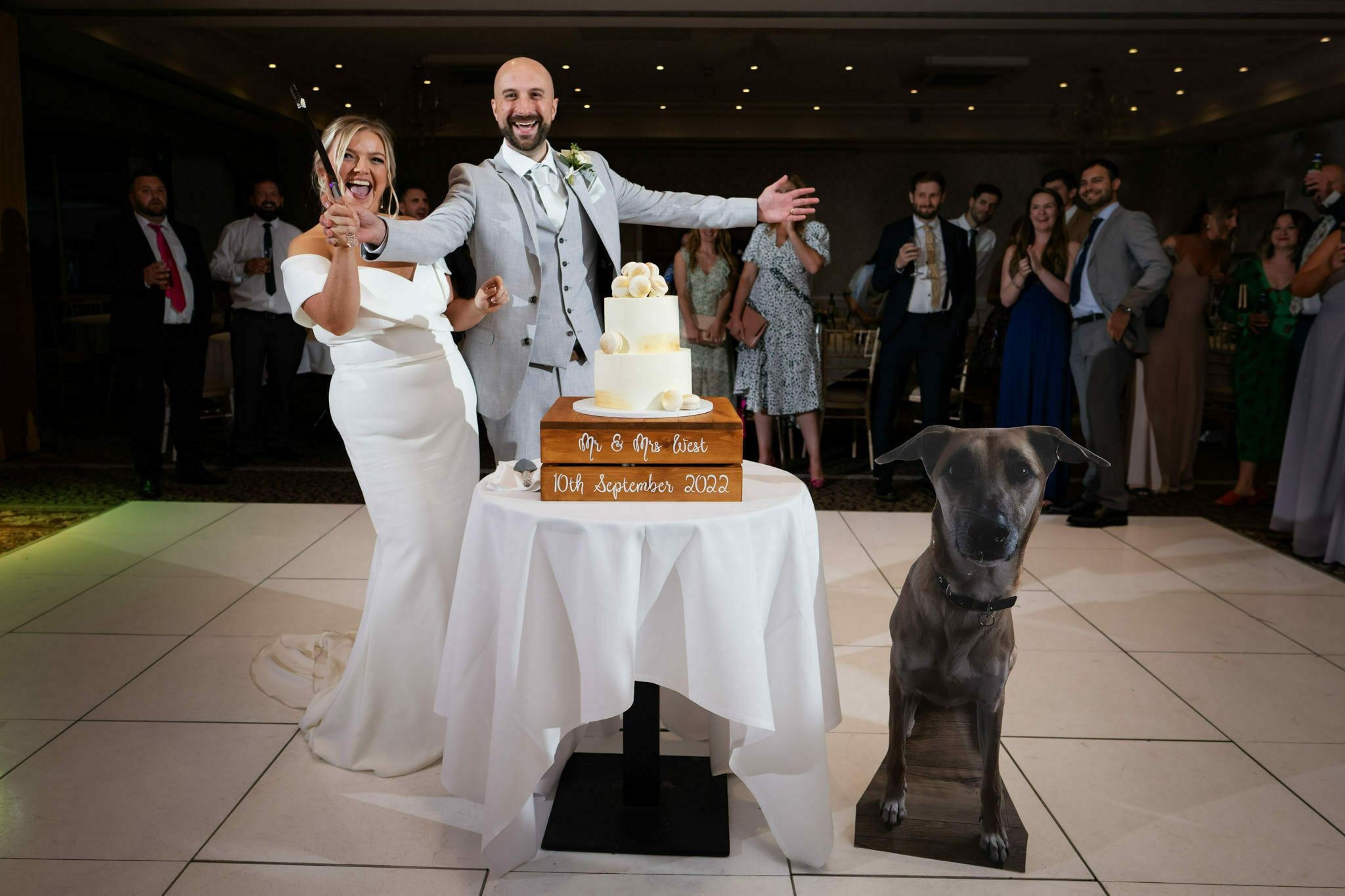 Cutting the cake at a Shendish Manor wedding the bride and groom give a big cheer, stood next to the cake table and a large cardboard cutout of their pet dog.