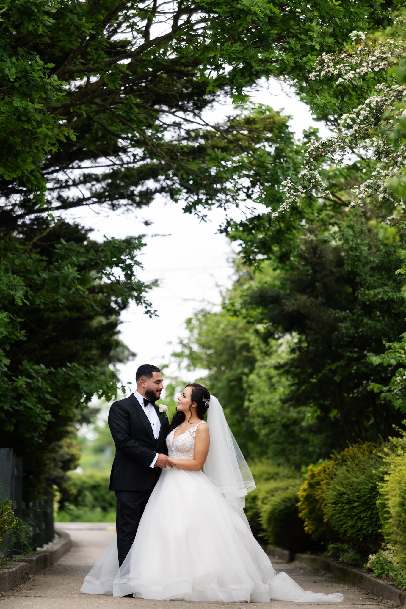A bride and groom stand in a garden at Friern Manor, looking at each other and smiling. The bride wears a white gown and a veil, while the groom is in a black tuxedo. Green foliage surrounds them.