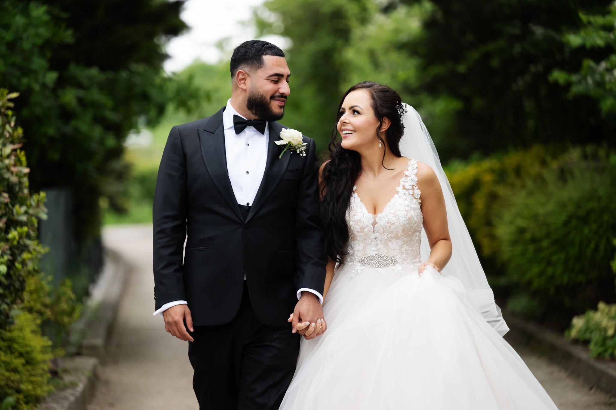 A couple smiles and holds hands while walking on a path in a garden at Friern Manor. The man is dressed in a black suit and bow tie, and the woman is in a white wedding gown and veil, celebrating their enchanting wedding day.