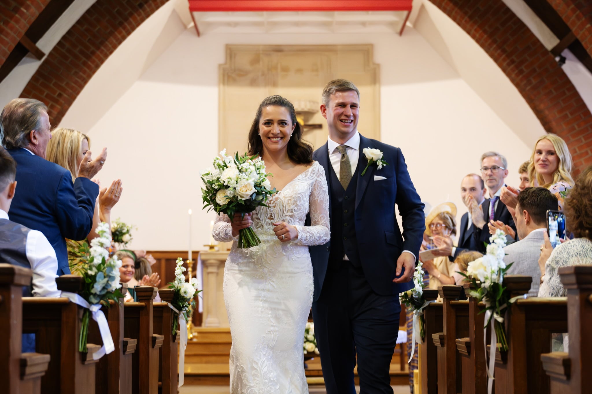 Charlotte and Matt's wedding at St Mary and St Joseph Church in Boxmoor was a heartwarming spectacle, as the bride in a white dress and the groom in a blue suit walked down the aisle holding flowers, while guests seated in pews applauded.