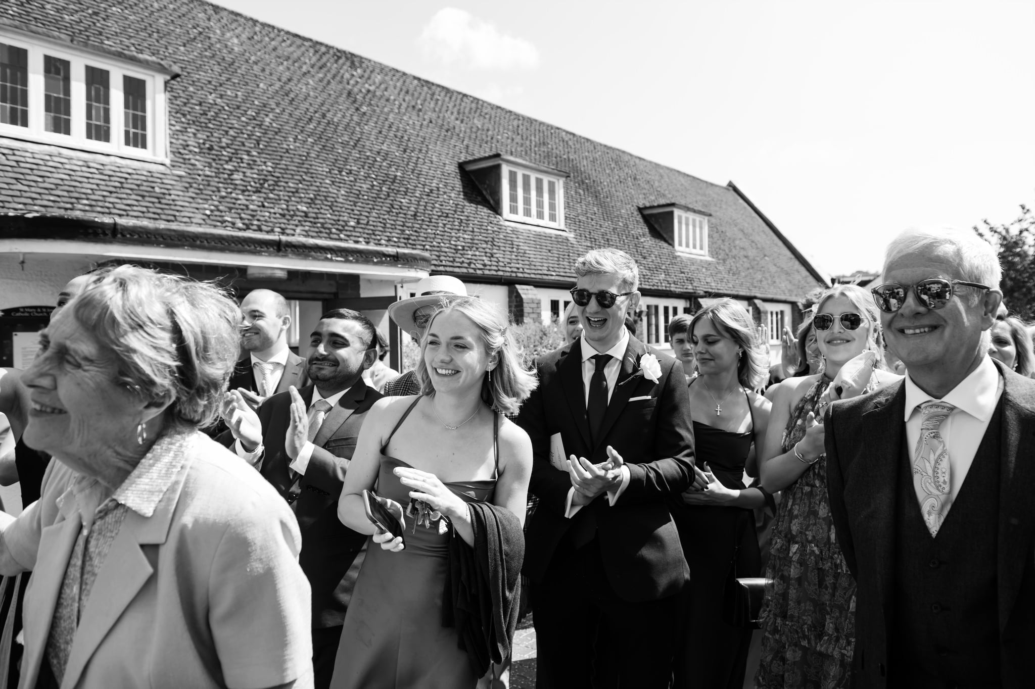A group of people, dressed in formal attire, are standing outdoors, smiling and applauding. The background shows a building with a sloped roof. The image is in black and white, capturing the joyous moments of Charlotte and Matt's wedding at St Mary and St Joseph Church in Boxmoor.