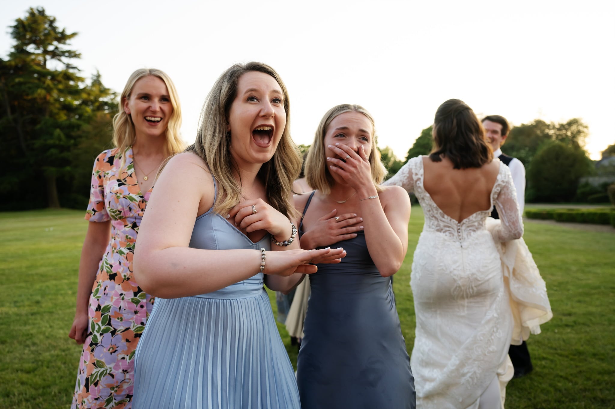 Four women are standing on a grassy area in the park at Charlotte & Matt's wedding. Two are dressed in blue dresses, looking surprised or excited. In the background, a bride and groom are seen embracing at Shendish Manor Wedding in Hemel Hempstead.