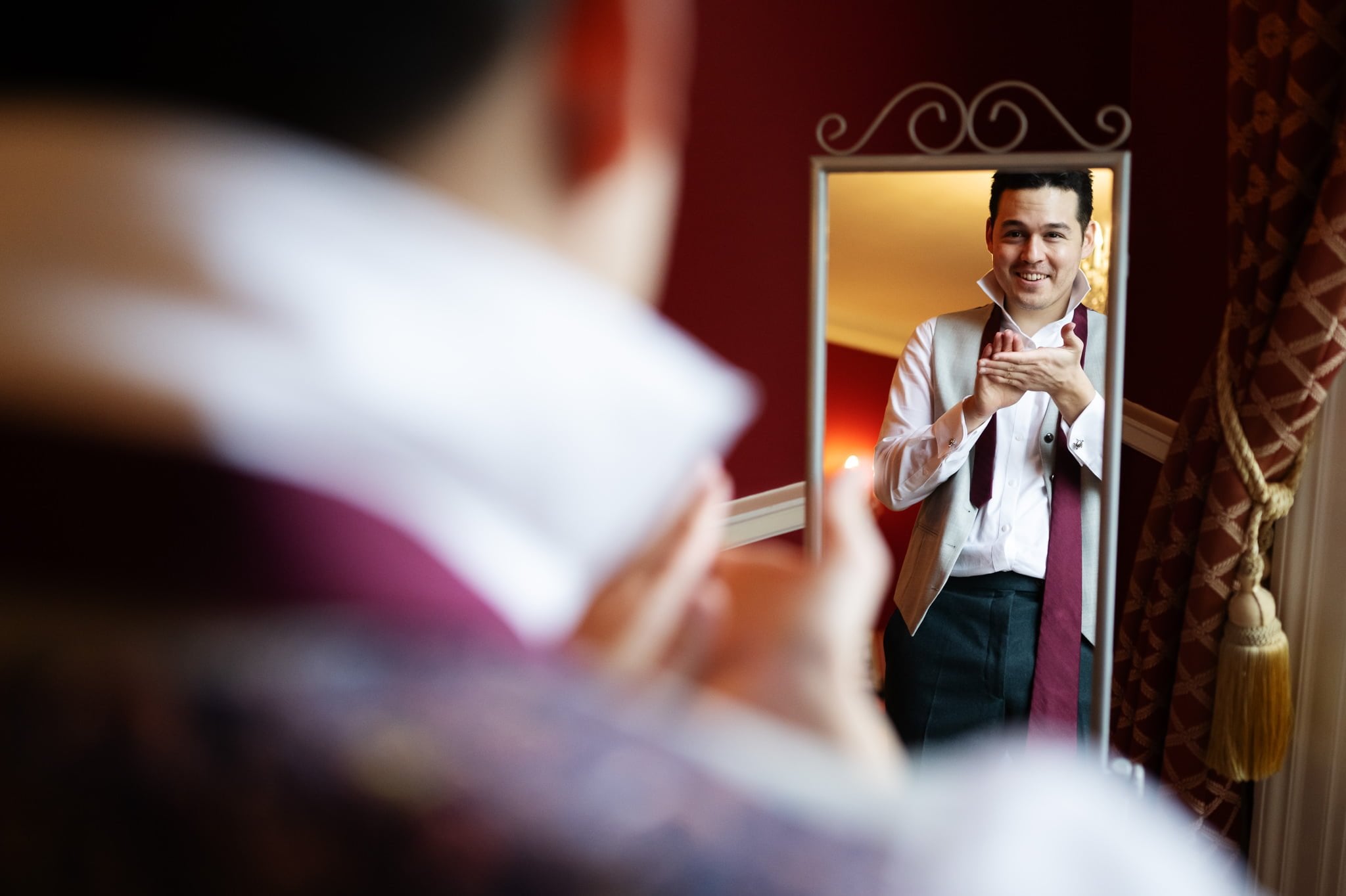 A man in formal attire is seen adjusting his bow tie in front of a mirror, smiling, as he gets ready for an Addington Palace wedding photography session.