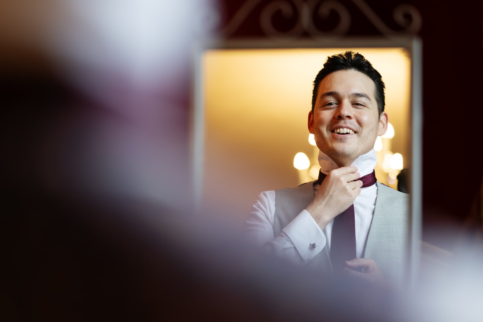 A man in a suit adjusts his tie while smiling at his reflection in the mirror, radiating confidence and joy—perfectly capturing the essence of Addington Palace wedding photography.