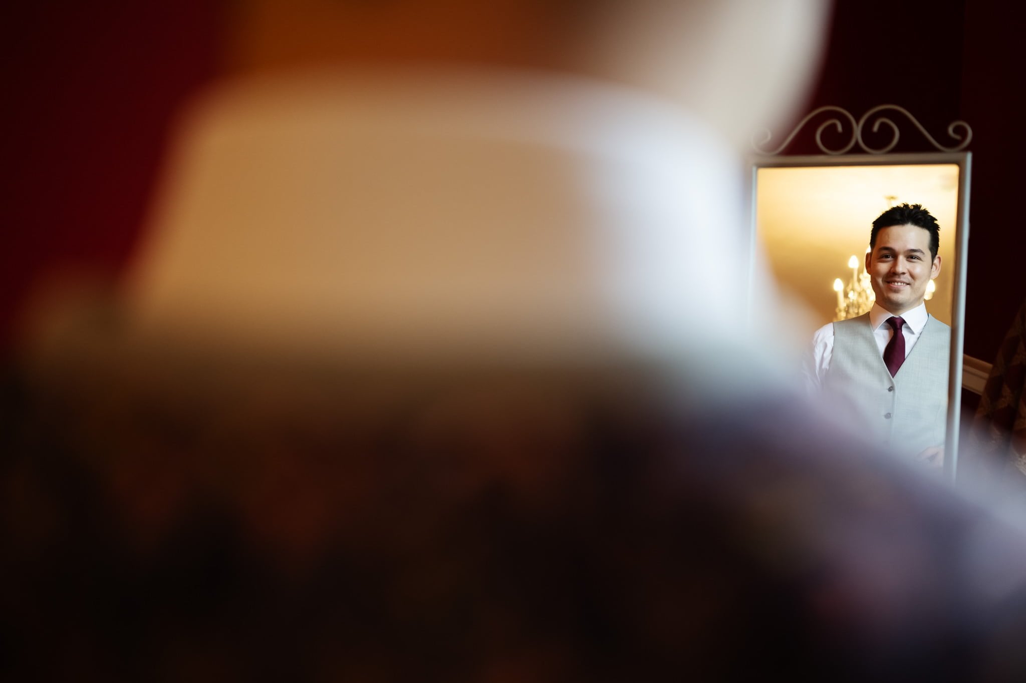 A person in formal attire is reflected in a mirror, with another person blurred in the foreground—a perfect scene for Addington Palace wedding photography.