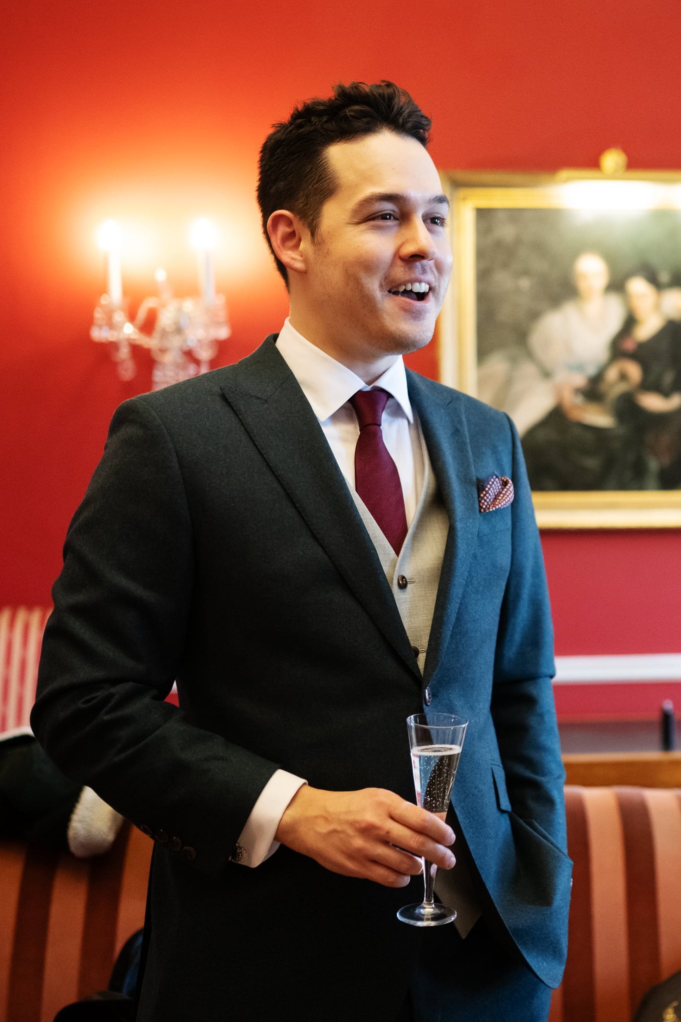 A man in a suit and tie is standing and holding a glass of champagne in a warmly lit room with red walls, a painting, and a chandelier in the background. This elegant scene could easily be part of an Addington Palace wedding, known for its stunning Surrey location.