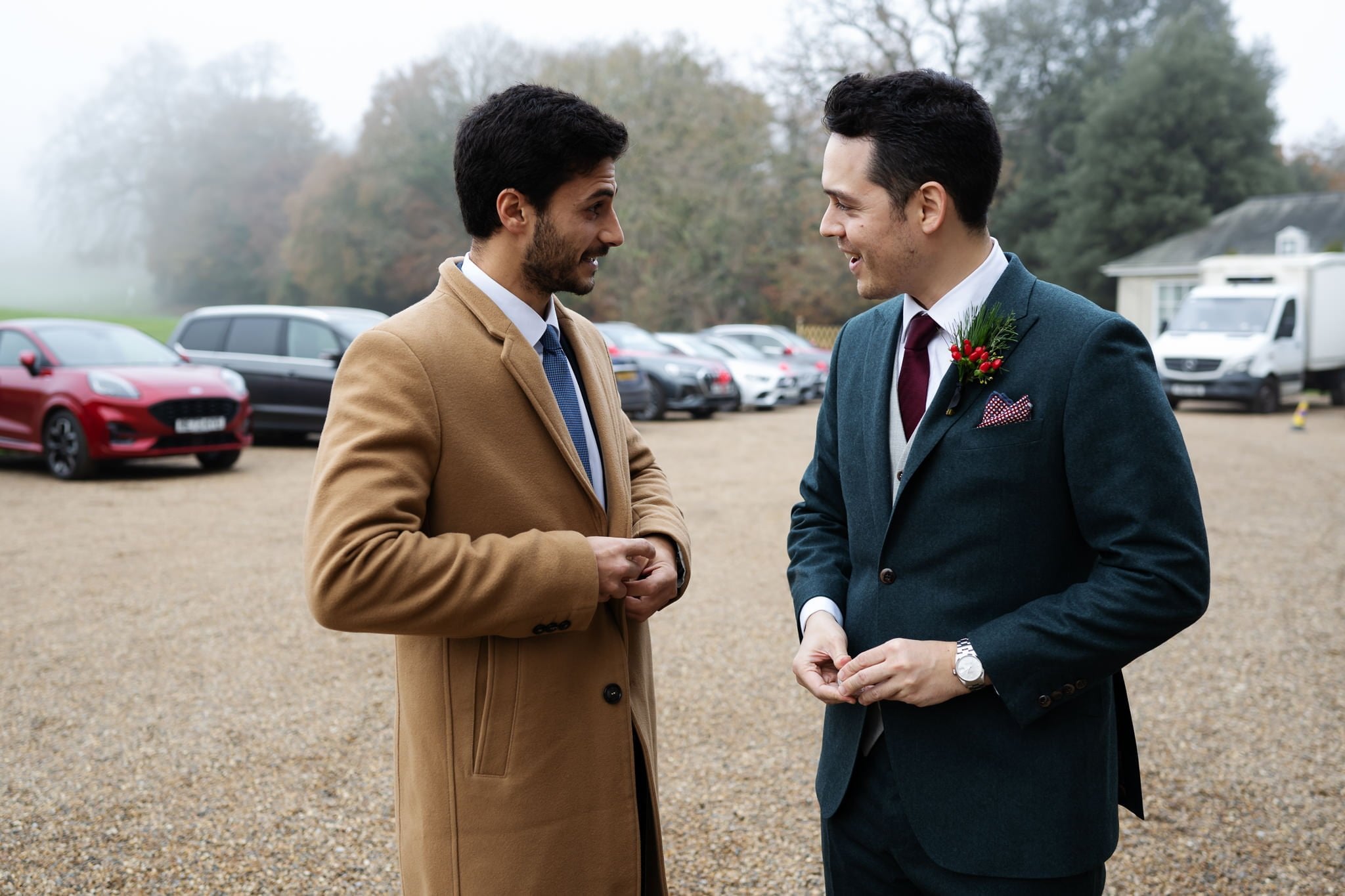 Two men in formal attire stand outside Addington Palace, engaging in conversation. One wears a camel coat and a blue tie, the other a green suit with a boutonniere and red tie. Cars and trees are in the background, capturing the charm of Surrey as seen by an Addington Palace wedding photographer.