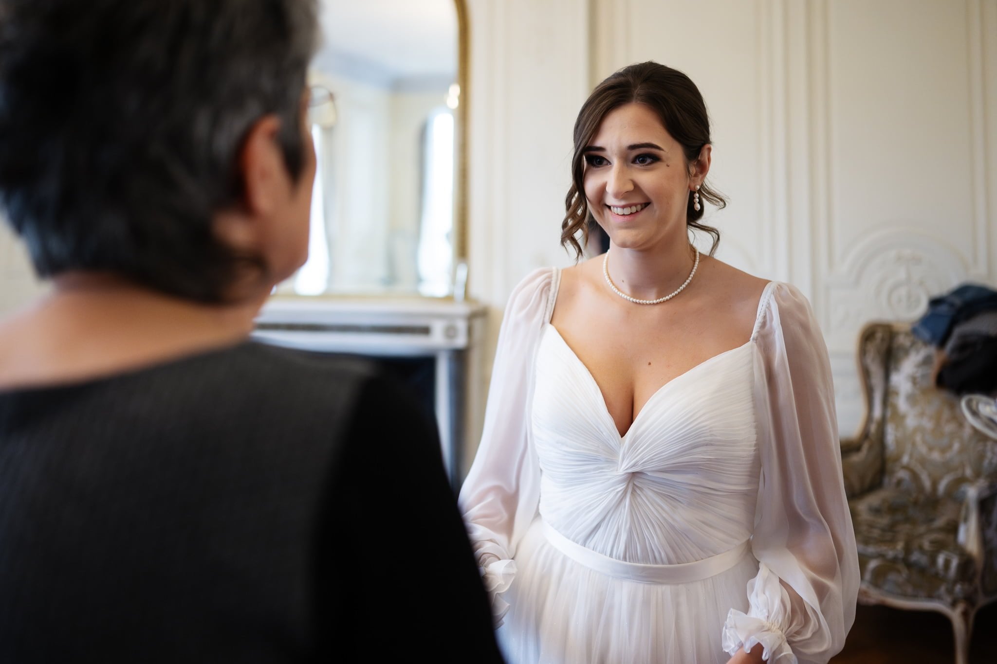 A woman in a white dress with a pearl necklace smiles at an older person in a black outfit inside a room with elegant decorations, perfectly capturing the charm of an Addington Palace wedding.
