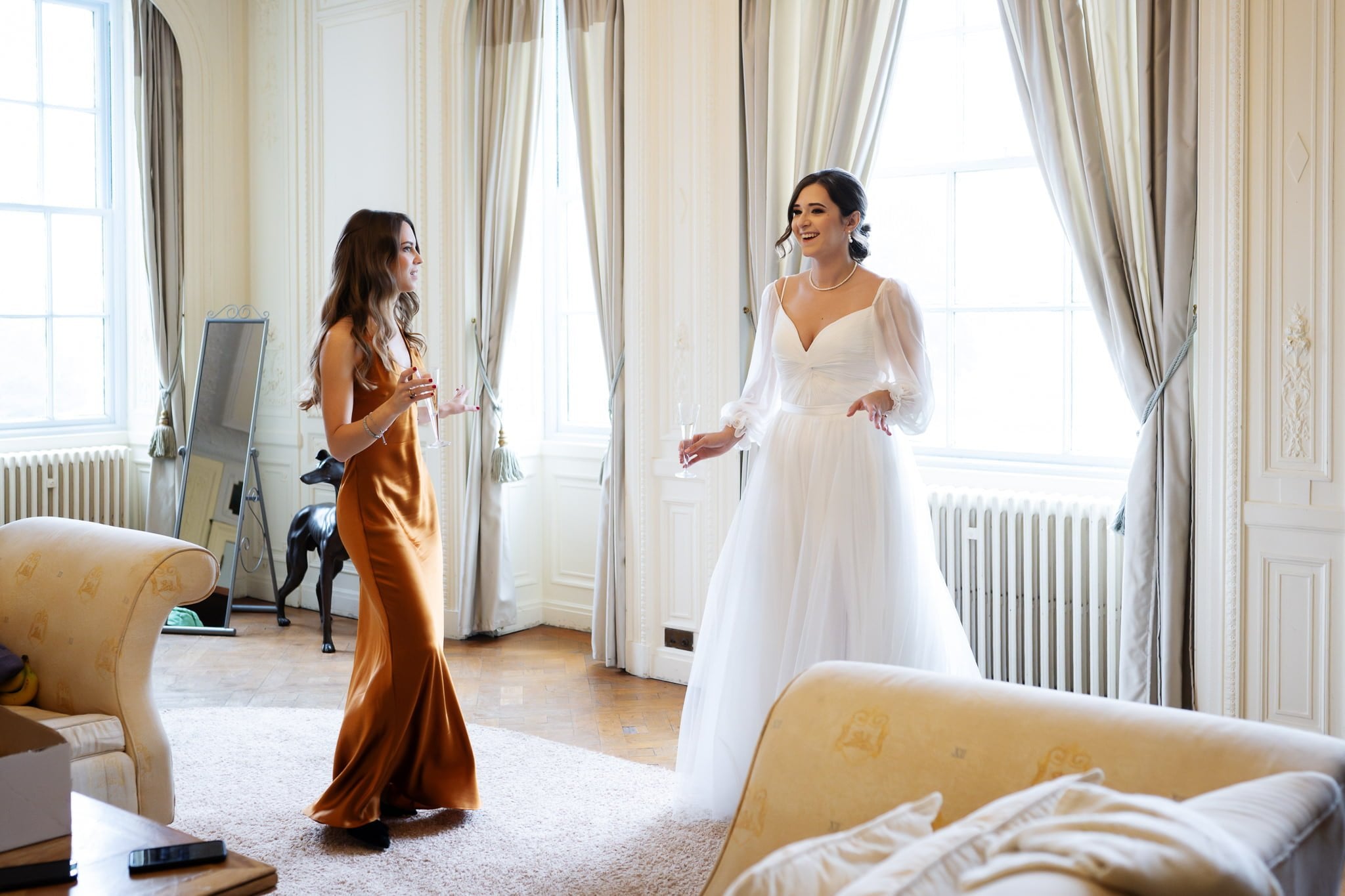 Two women are having a conversation in a well-lit room with large windows at Addington Palace. One is wearing a white gown and the other a brown dress. They stand near sofas and a radiator, perfectly capturing the elegance typical of Surrey's renowned wedding venues.