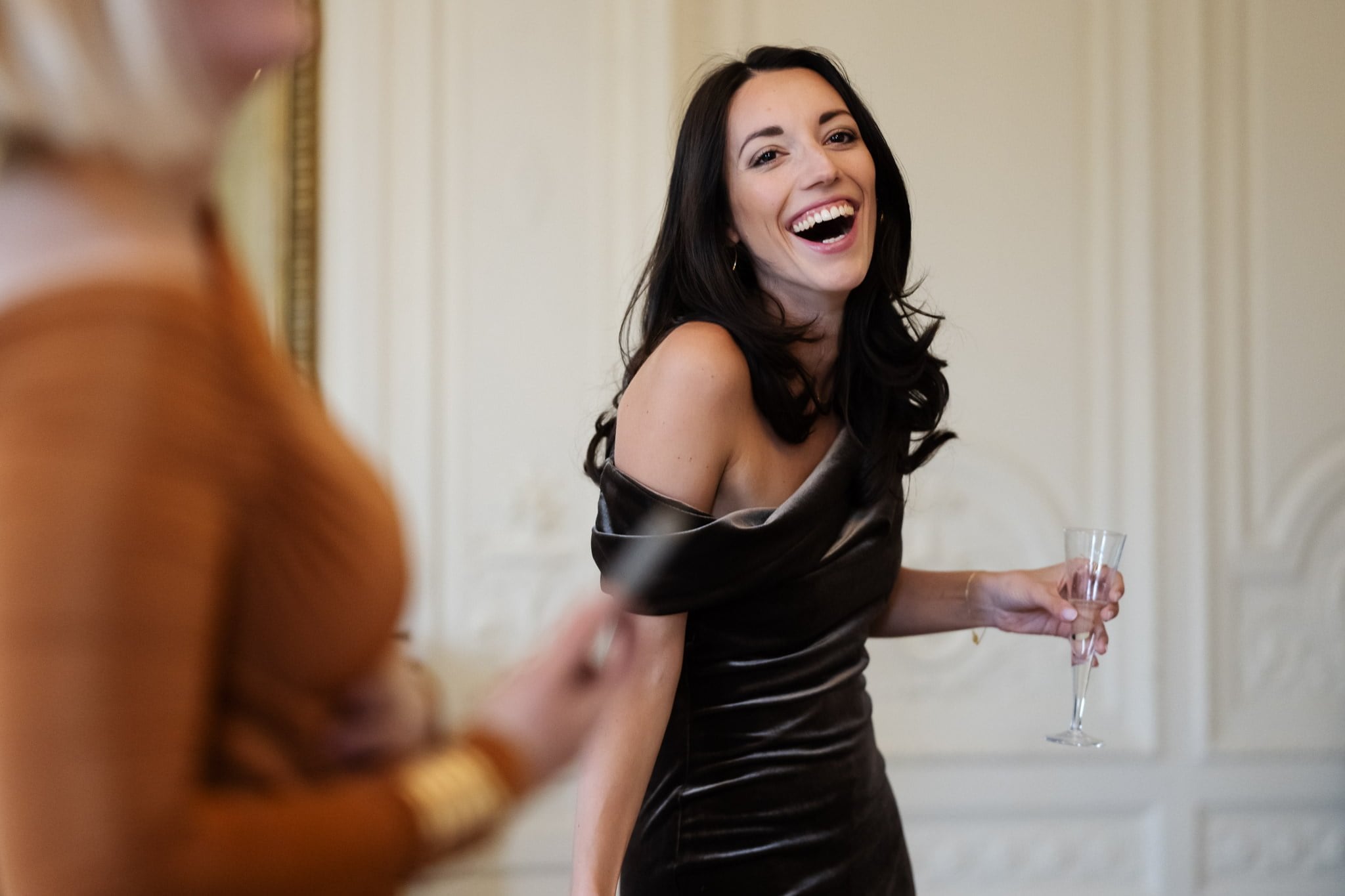 Two women are having a conversation at an event. The woman in the foreground, perhaps captured by an Addington Palace wedding photographer, is wearing a brown dress and holding a champagne flute, while the woman in the background is laughing.