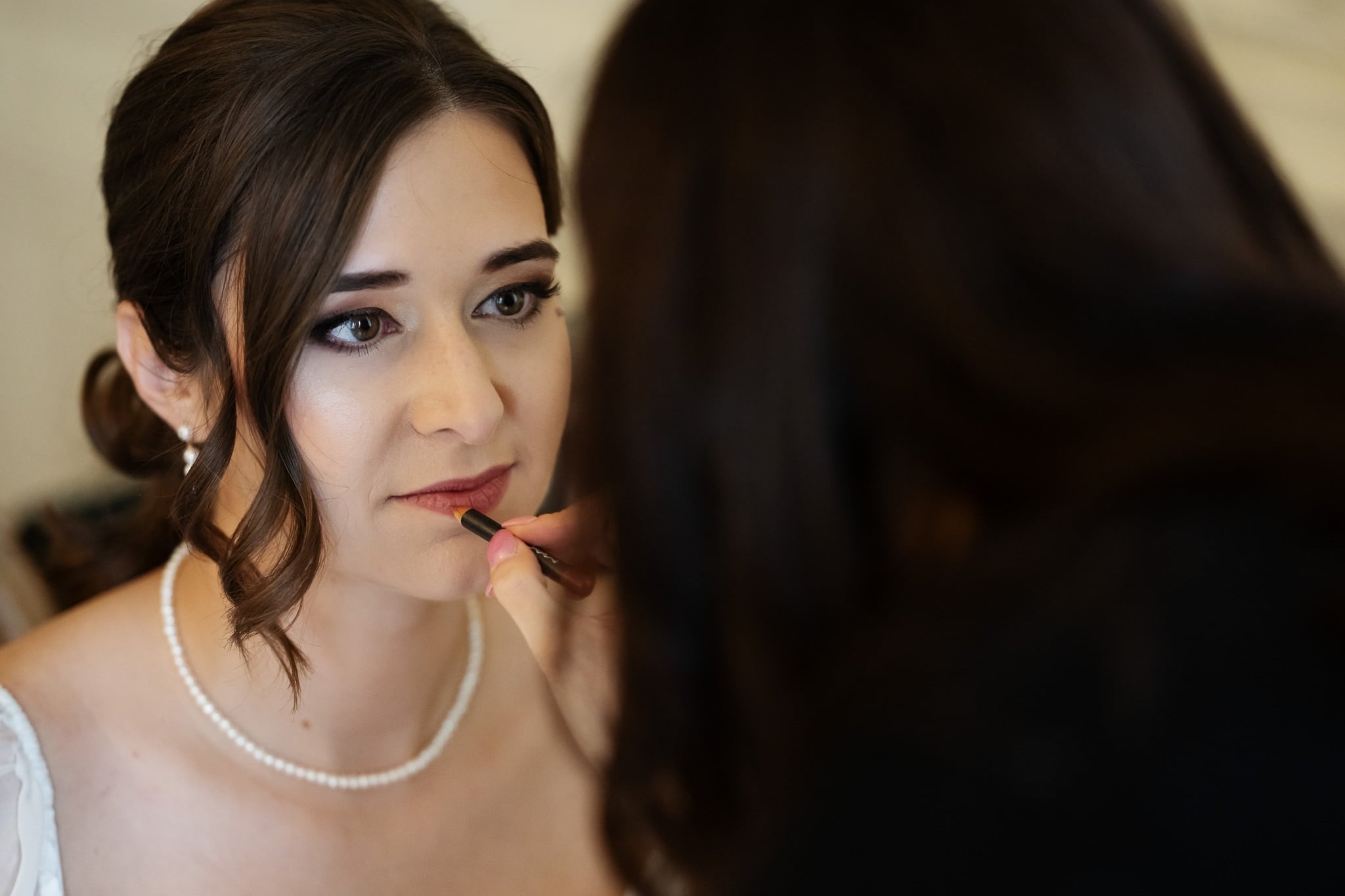 A woman is having her makeup applied by another person, focusing on her lips. She is wearing a white dress and a pearl necklace, as an Addington Palace wedding photographer captures the intimate moment before the ceremony.