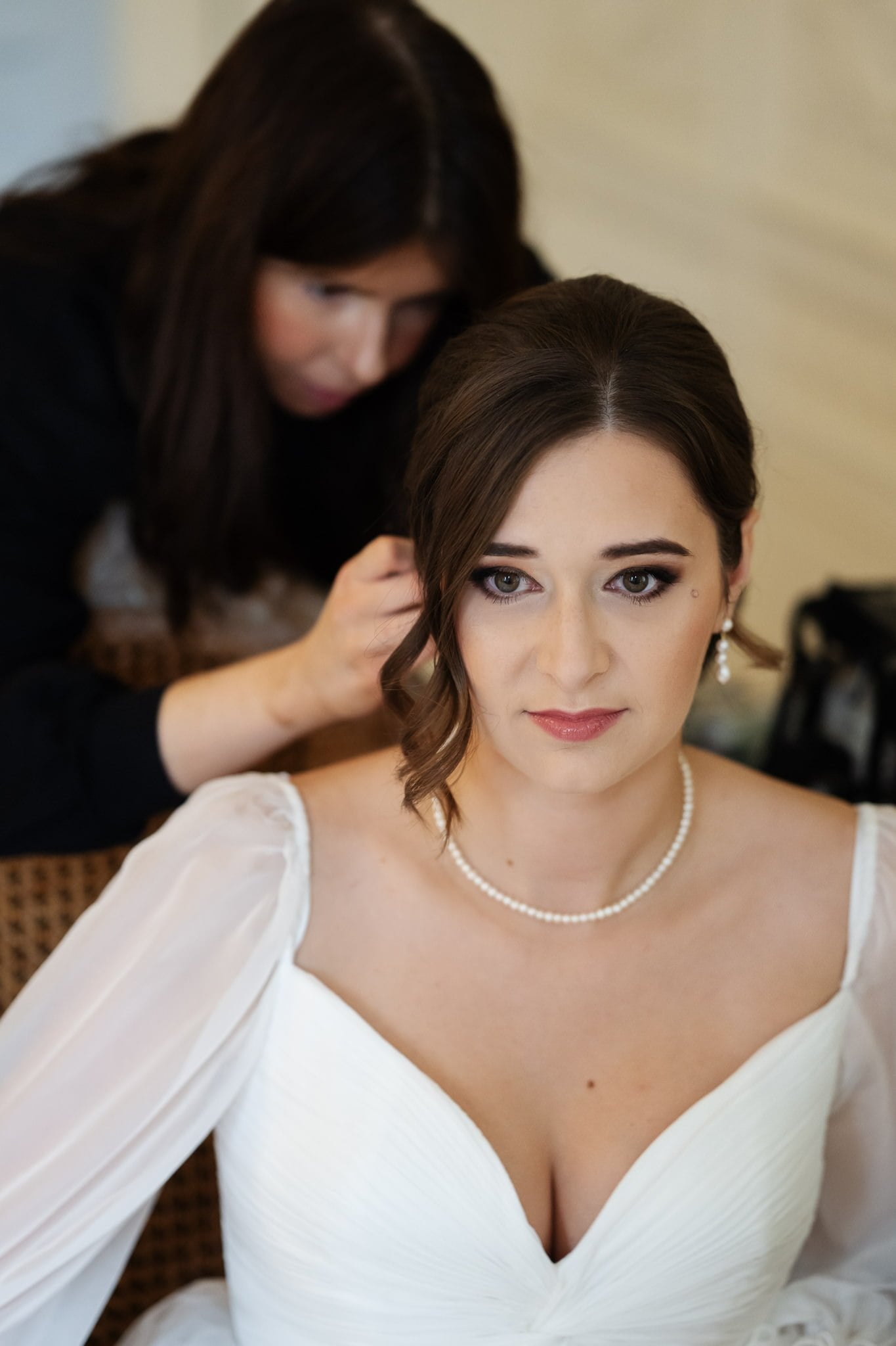 A bride in a white dress and pearl necklace has her hair styled by a person in the background, ready for her Addington Palace wedding in Surrey.