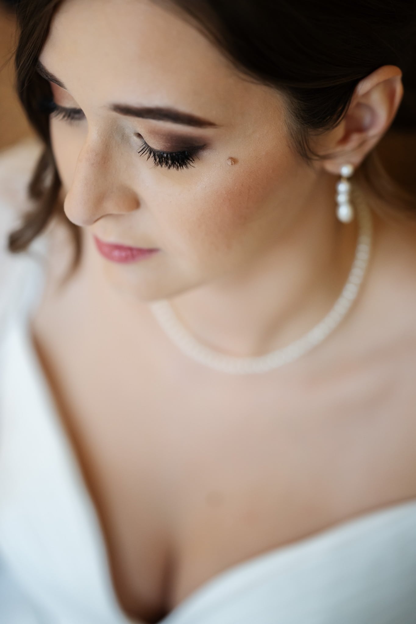 Close-up of a woman with long eyelashes and wearing a pearl necklace and earrings. She appears to be looking downward, perhaps lost in thought during her wedding at Addington Palace in Surrey.