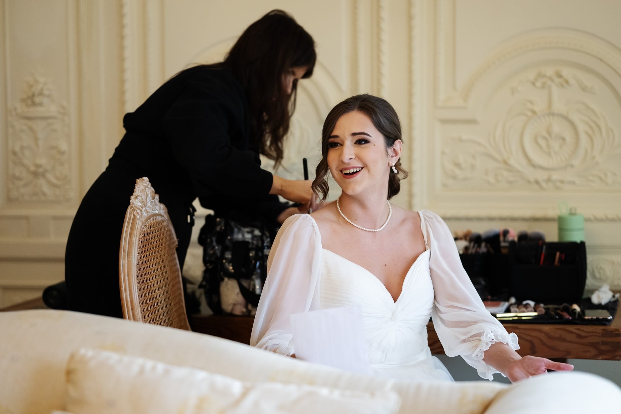 In an ornate room, a bride in a white dress and pearl necklace sits smiling as a makeup artist works at the table, embodying the elegance captured by Addington Palace wedding photography.