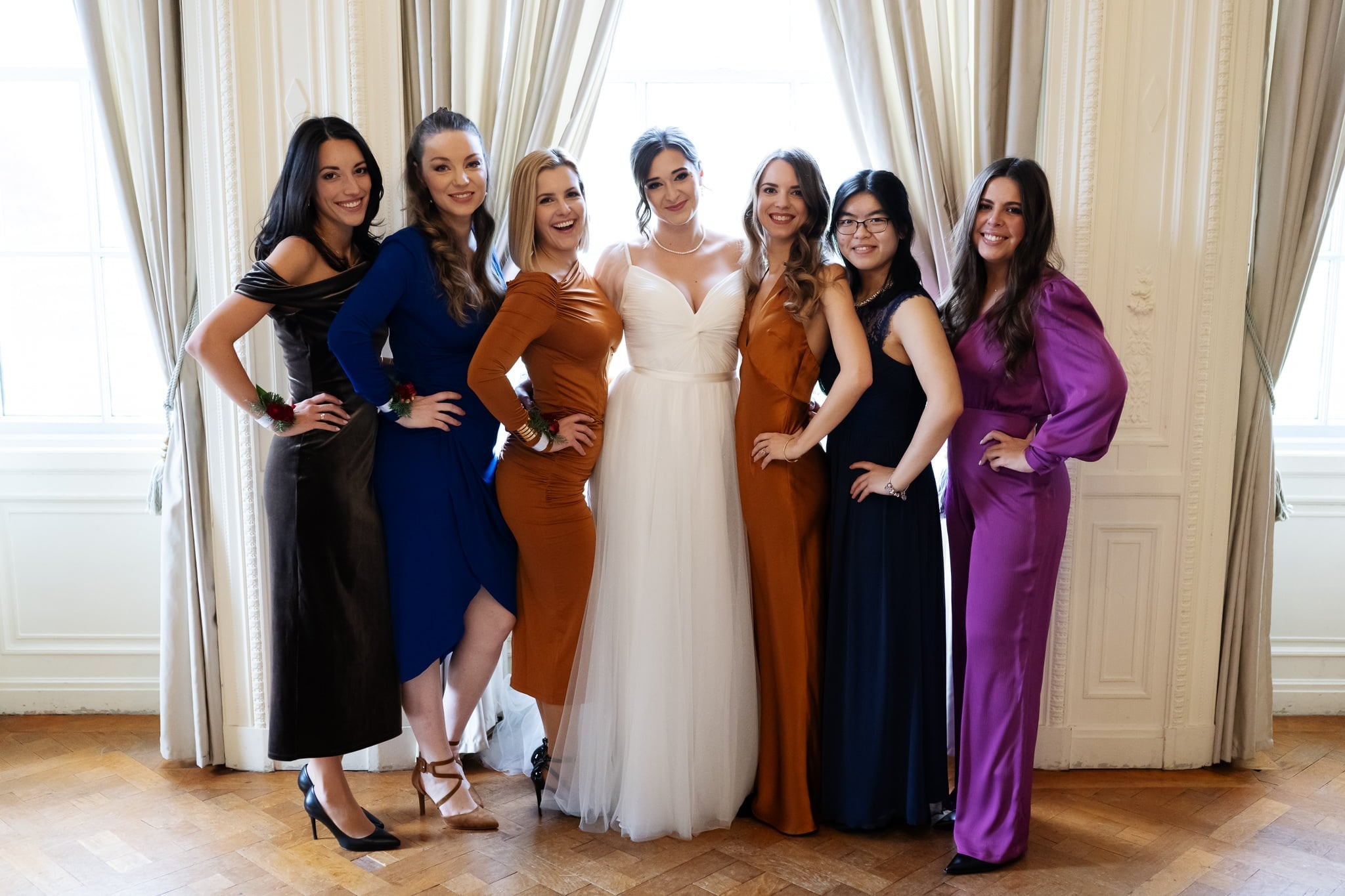 A bride in a white dress poses with six women in colorful dresses, standing in front of large windows with beige curtains at an enchanting Addington Palace wedding.