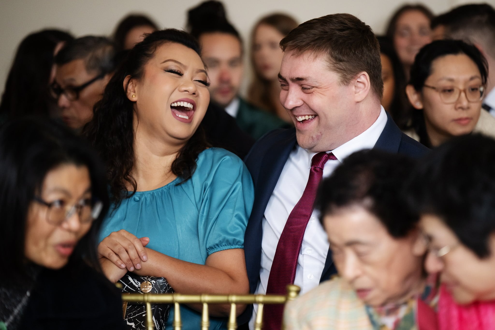 Two people in formal attire are seated close together, laughing among a larger, more serious audience at an Addington Palace wedding.