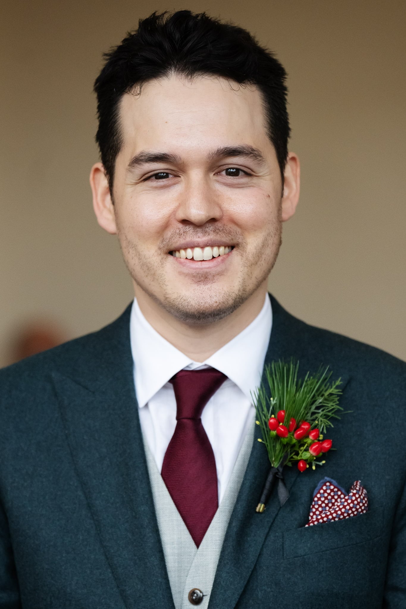 A man in a green suit, white shirt, and red tie is smiling. He wears a red and white polka dot pocket square and a boutonniere with green foliage and red berries, looking picture-perfect for an Addington Palace wedding in Surrey.