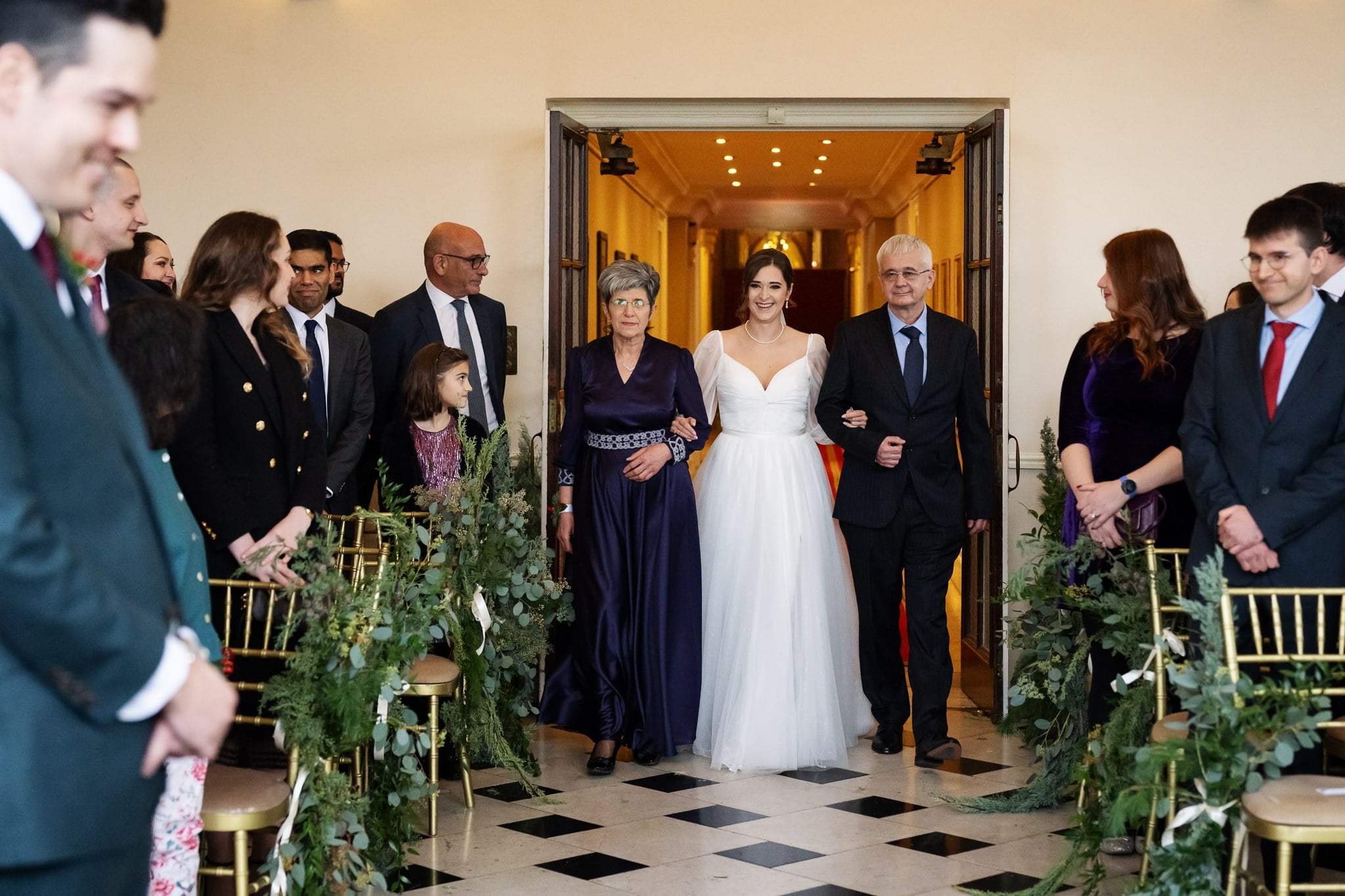 A bride in a white gown walks down an aisle at Addington Palace, flanked by two older adults in formal attire, with seated guests looking on. This Surrey venue provides the perfect backdrop for your wedding photography, capturing every moment beautifully.