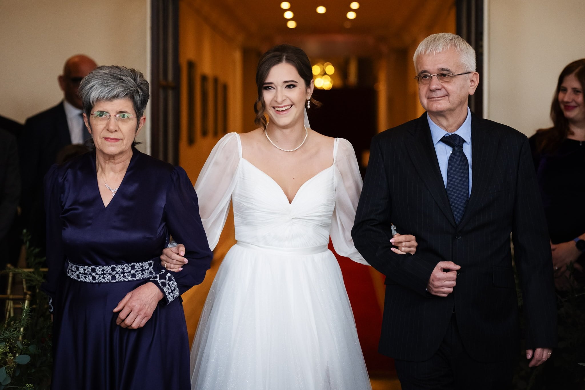 A bride in a white gown is smiling and standing between an elderly woman in a purple dress and an elderly man in a dark suit, holding their arms, during an Addington Palace wedding, with framed pictures adorning the corridor walls.