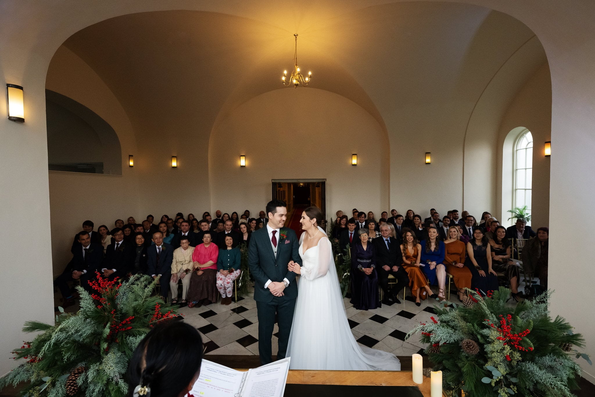 A bride and groom stand at the altar in a spacious, arch-ceilinged venue, facing an audience of seated guests. The room, perfect for their Addington Palace wedding in Surrey, is decorated with greenery and red berries, and lit by wall-mounted lamps.