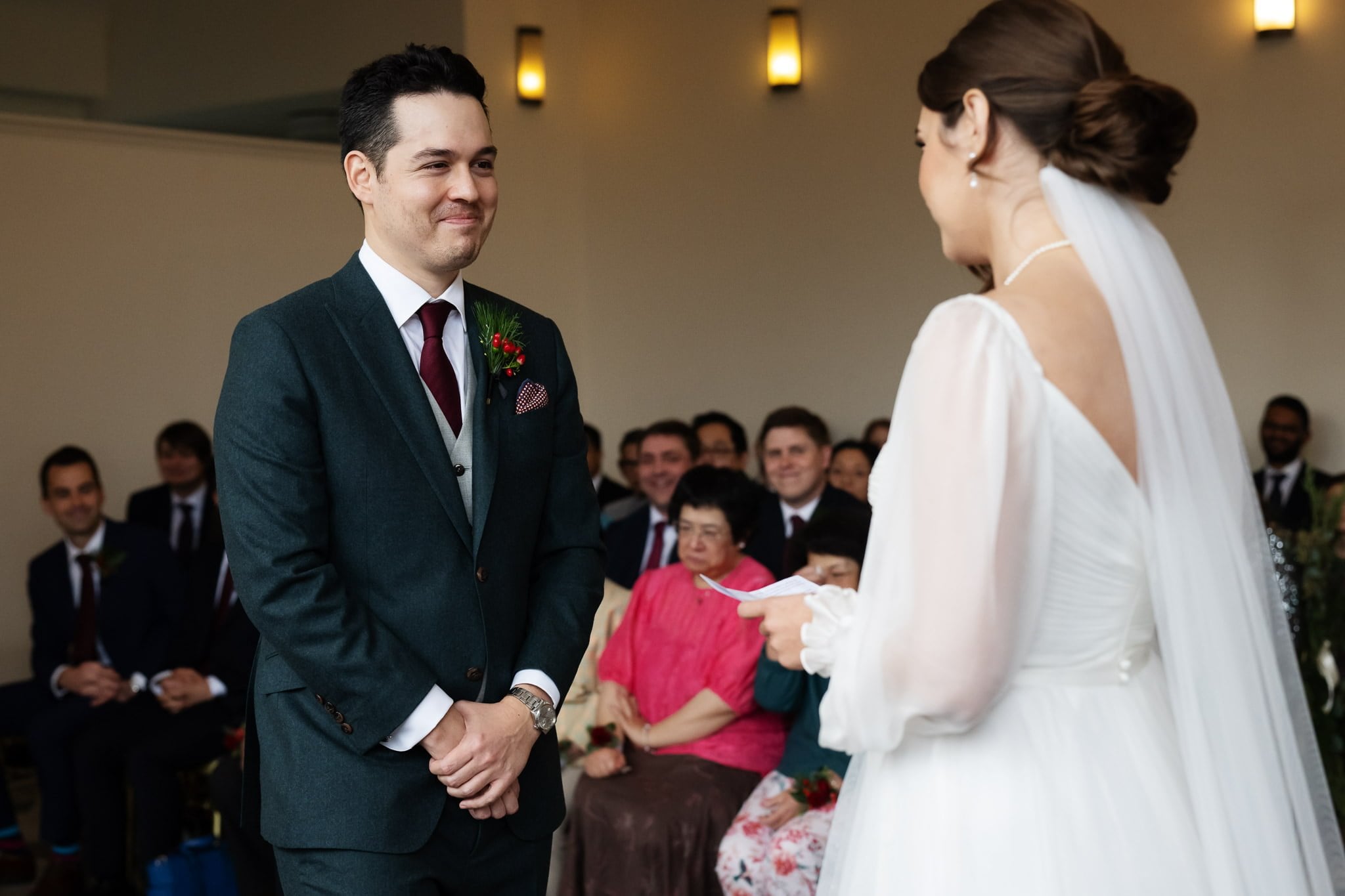 A bride and groom stand facing each other during their Addington Palace wedding ceremony in Surrey, with guests seated and watching them in the background. The bride holds a piece of paper, possibly reading her vows.