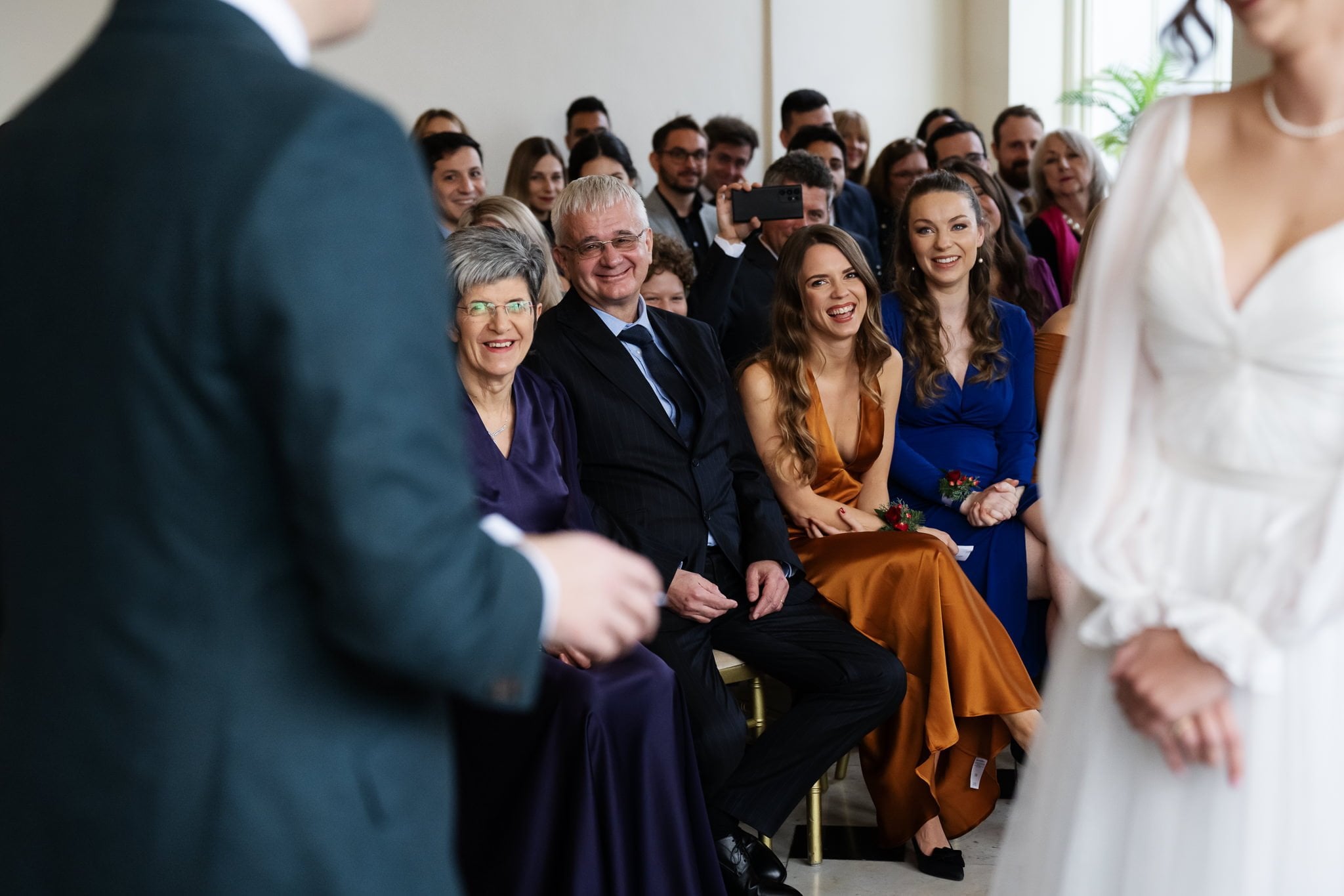 A group of people, including an older couple and two younger women in colorful dresses, sit among a larger audience smiling at two individuals in formal attire standing in the foreground. The joyful scene captures the essence of an Addington Palace wedding.