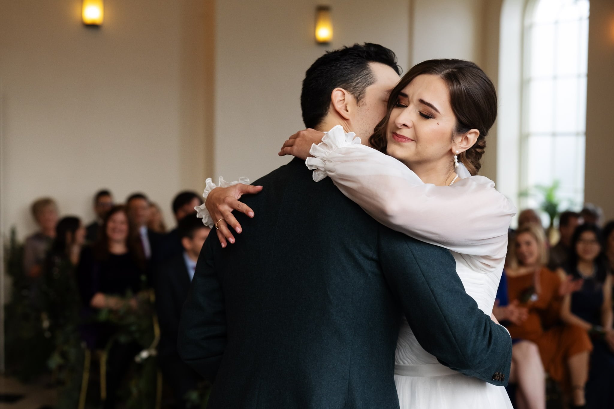 A couple shares an embrace at their Addington Palace wedding, with guests seated in the background inside a well-lit room in Surrey.