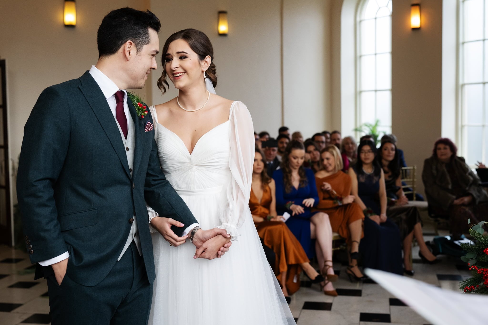 A bride and groom stand holding hands and smiling at each other during their Addington Palace wedding, with seated guests in the background.