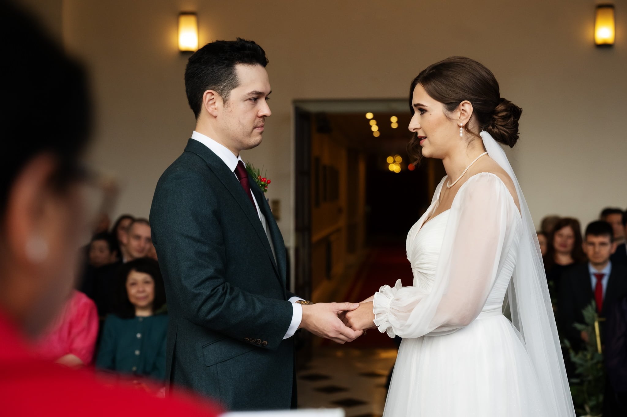 A bride and groom stand facing each other and holding hands during their wedding ceremony at Addington Palace, with guests seated in the background.