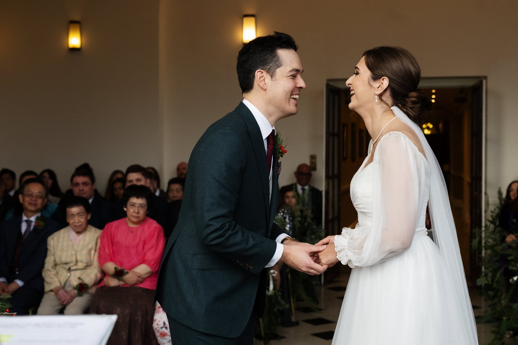 A couple is laughing and holding hands during their Addington Palace wedding ceremony while guests watch from their seats in the background, capturing a beautiful moment of Surrey wedding photography.