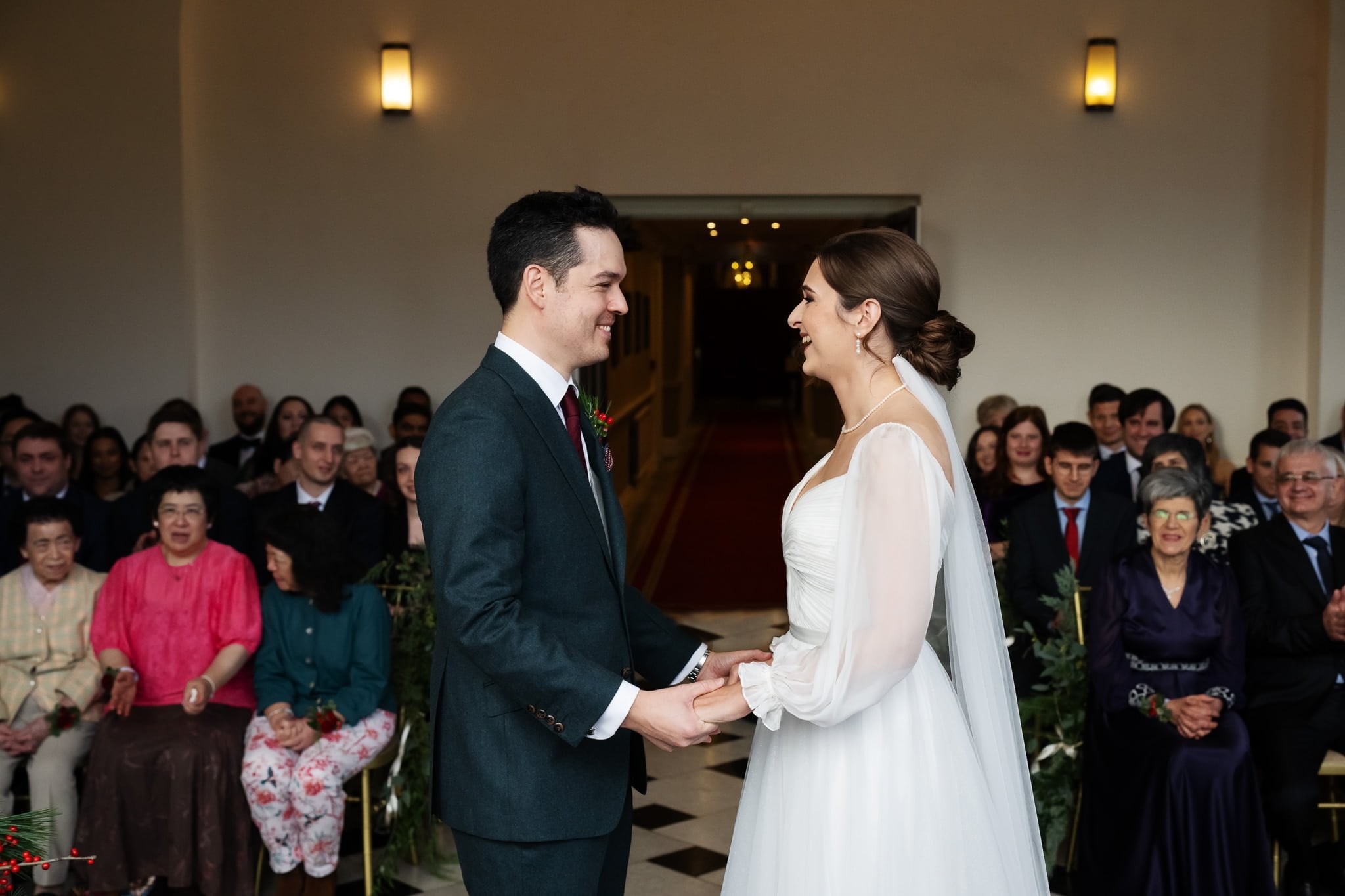 A bride and groom smile at each other, holding hands in a well-lit room at Addington Palace, surrounded by seated guests during their wedding ceremony.