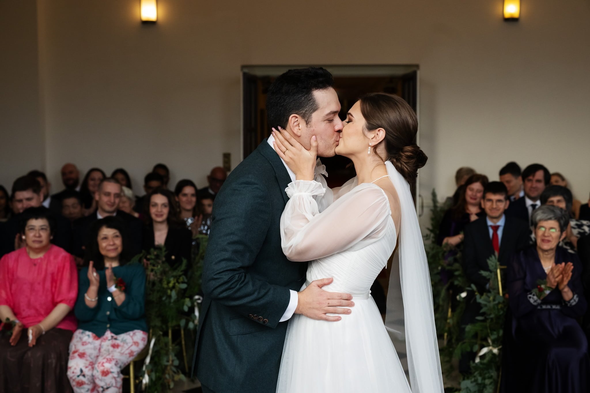 A bride in a white gown and a groom in a green suit kiss at their wedding ceremony. Guests are seated in the background, with some clapping. Captured by an Addington Palace wedding photographer, this perfect moment highlights the joy of their special day.