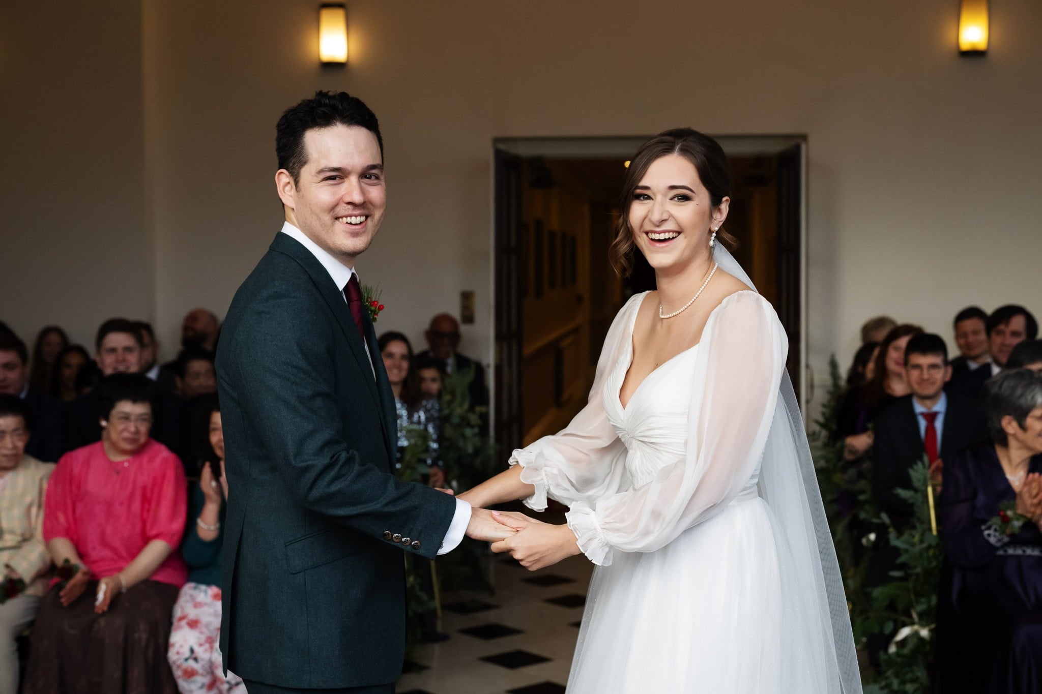 A couple holding hands and smiling at their wedding ceremony, with guests seated in the background—a perfect moment captured by Addington Palace wedding photography in Surrey.