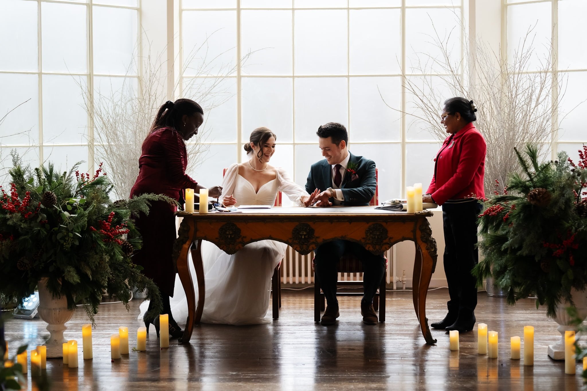 At an Addington Palace wedding, a bride and groom sit at a table signing documents, accompanied by two women standing on either side in a room decorated with greenery and candlelight.