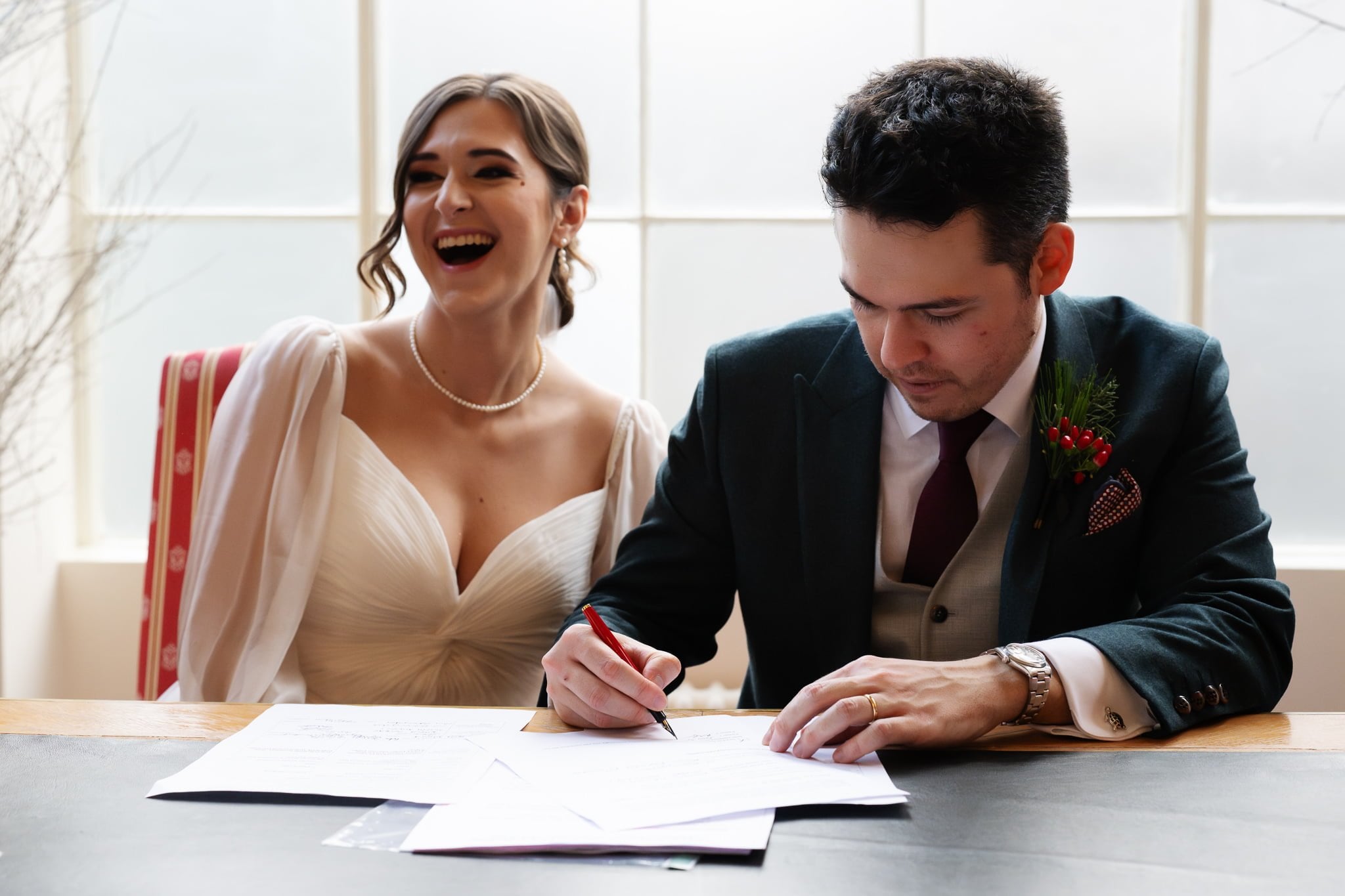 A bride and groom are seated at a table at their Addington Palace wedding. The groom is signing a document while the bride, clearly joyful, laughs heartily.