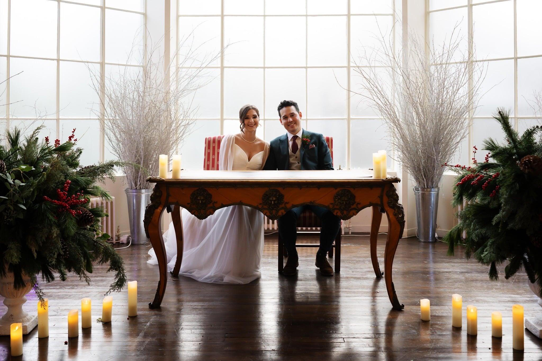 A bride and groom seated at an ornate wooden table, surrounded by festive greenery and candles, with large windows in the background—a timeless moment captured during their Addington Palace wedding.