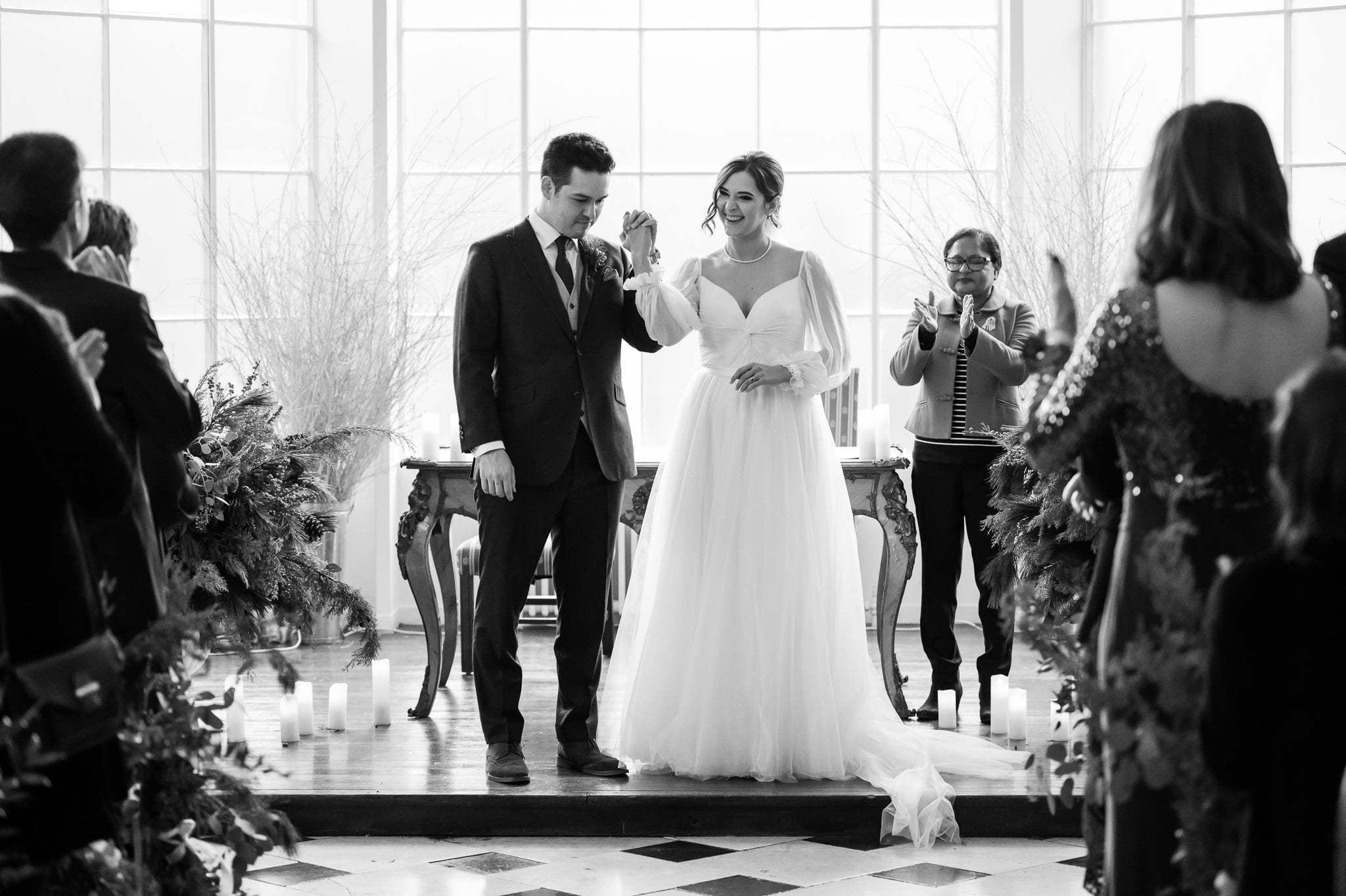 A couple, dressed in wedding attire, holds hands and stands at the altar surrounded by family and friends in a well-lit room with large windows, capturing the essence of an Addington Palace wedding.