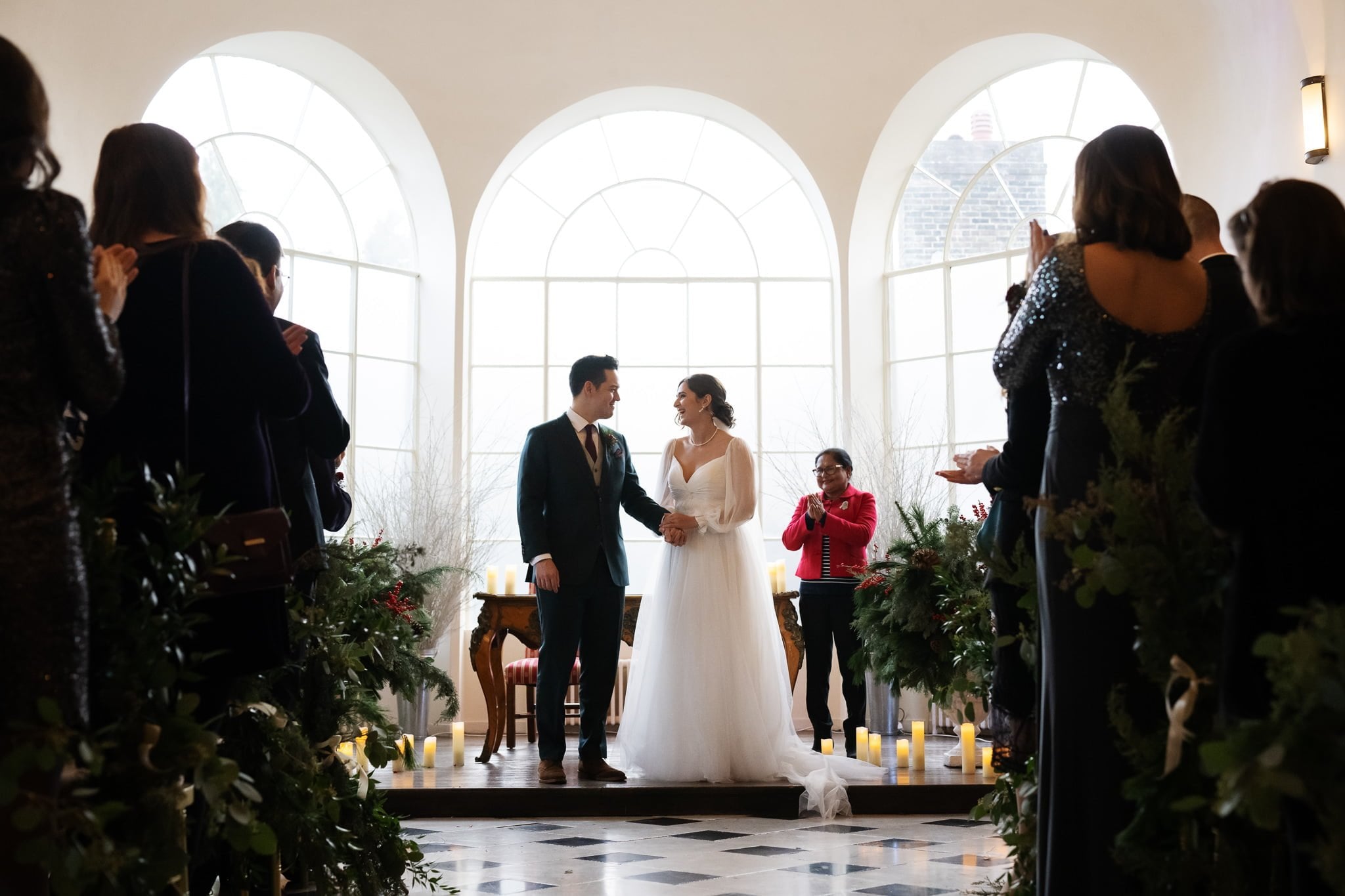 A bride and groom stand hand-in-hand at the altar, facing each other in a well-lit room with floor-to-ceiling windows at their Addington Palace wedding. Guests seated on either side are standing and applauding.