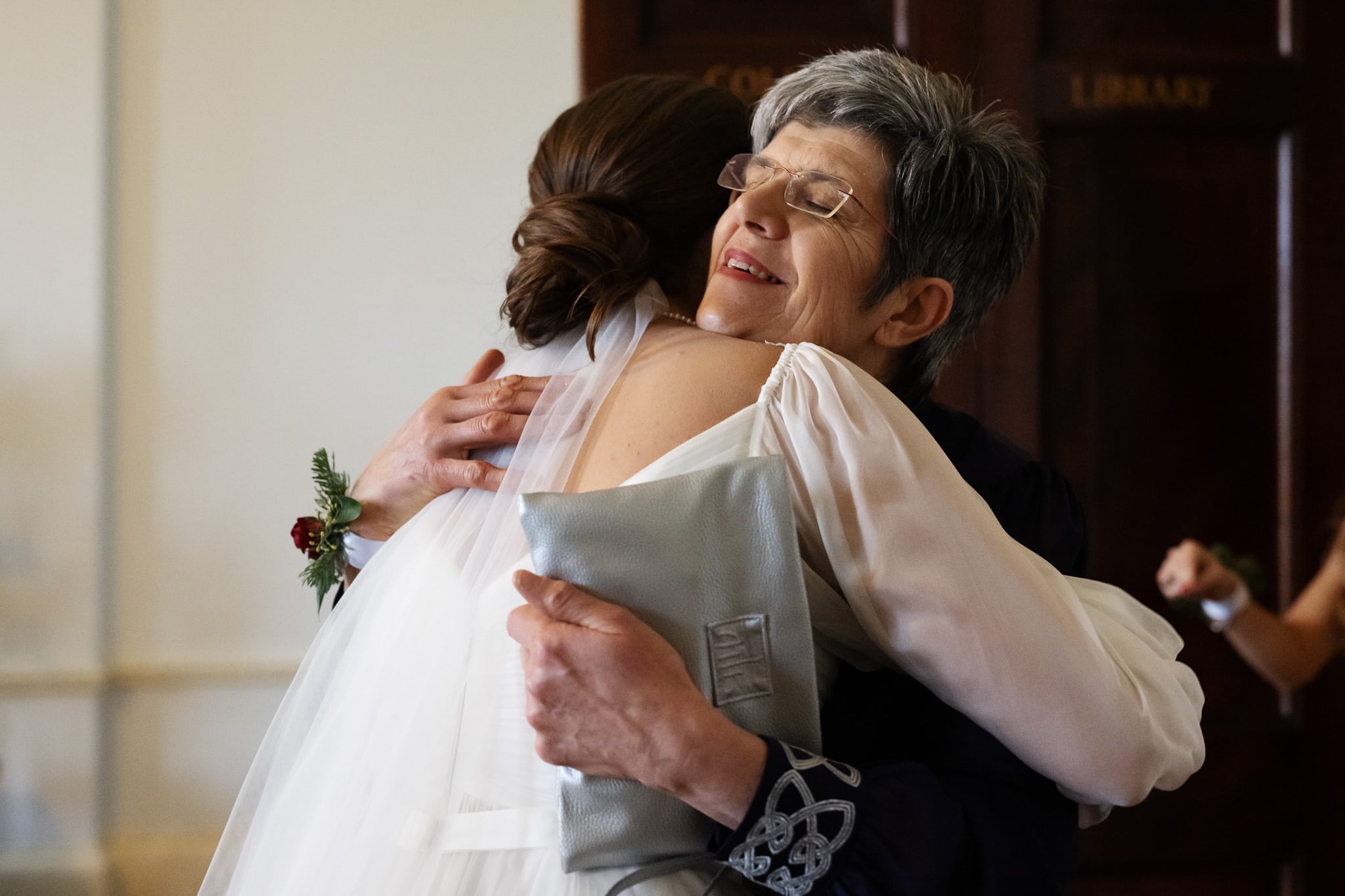 At an Addington Palace wedding, a bride in a white dress embraces an older woman with short gray hair, glasses, and a blue outfit. The older woman holds a gray clutch bag and smiles with eyes closed.
