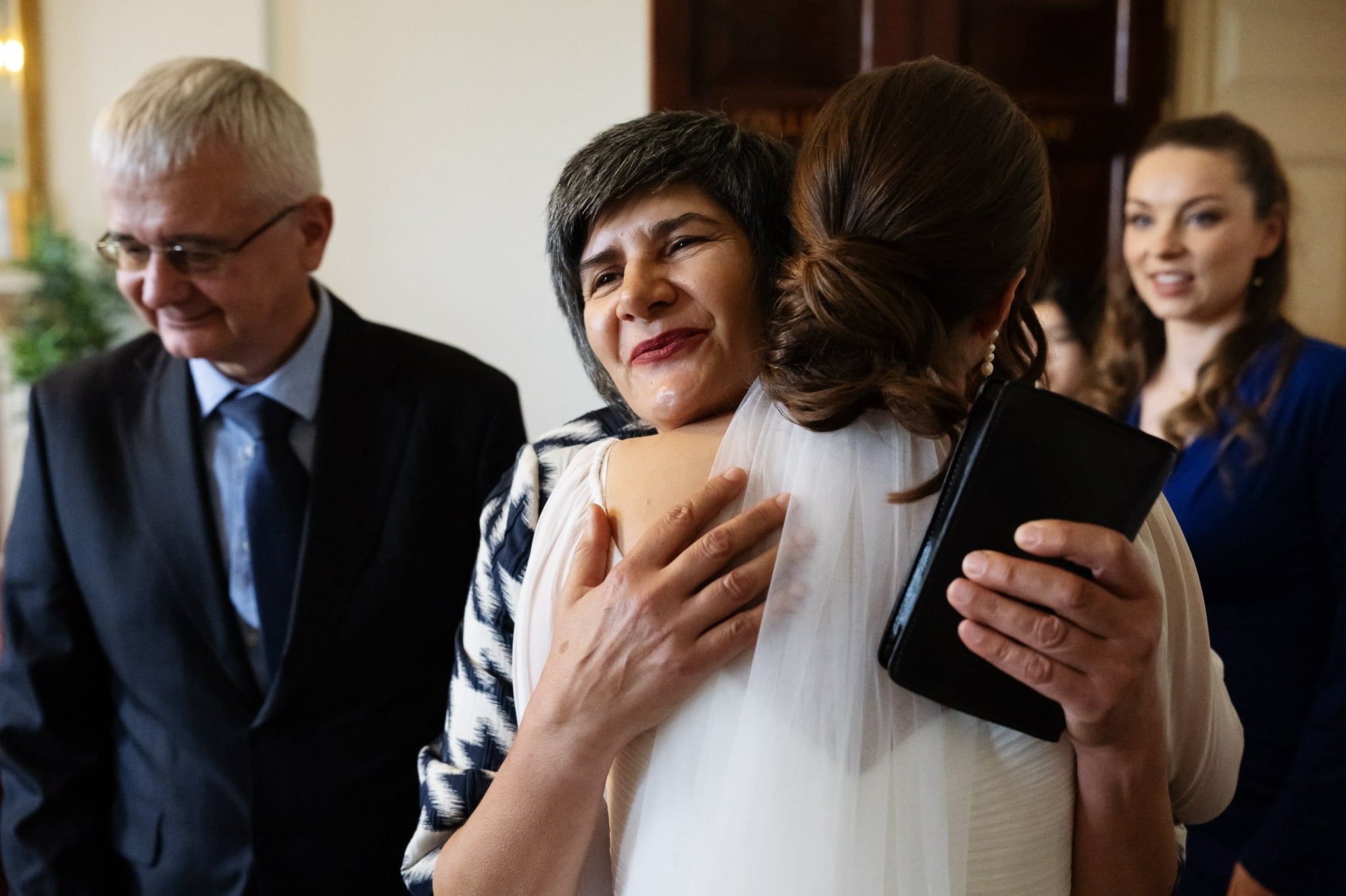 At an Addington Palace wedding, a woman in a veil hugs another who is holding a black wallet, while an older man and younger woman stand nearby, observing the moment.